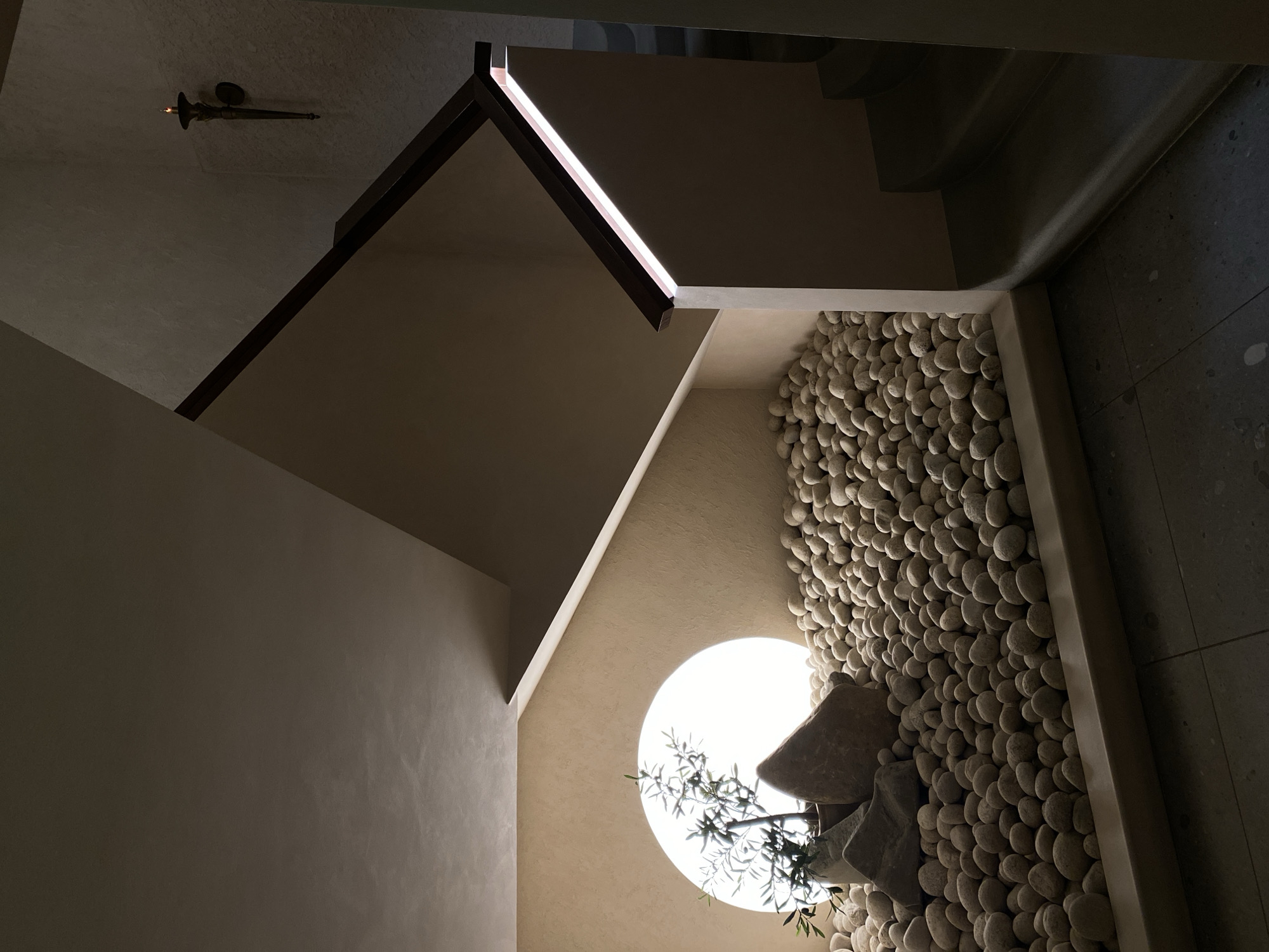 Captured from a lower vantage point in an architectural setting, the image features a confluence of textures, shapes, and light. Walls with a slightly textured surface converge around a circular window set into a wall covered with smooth, rounded stones. Inside, a shrub sits in front of the window. Above the window is a set of stairs. The top of the frame includes a lighting fixture, in front of textured walls.