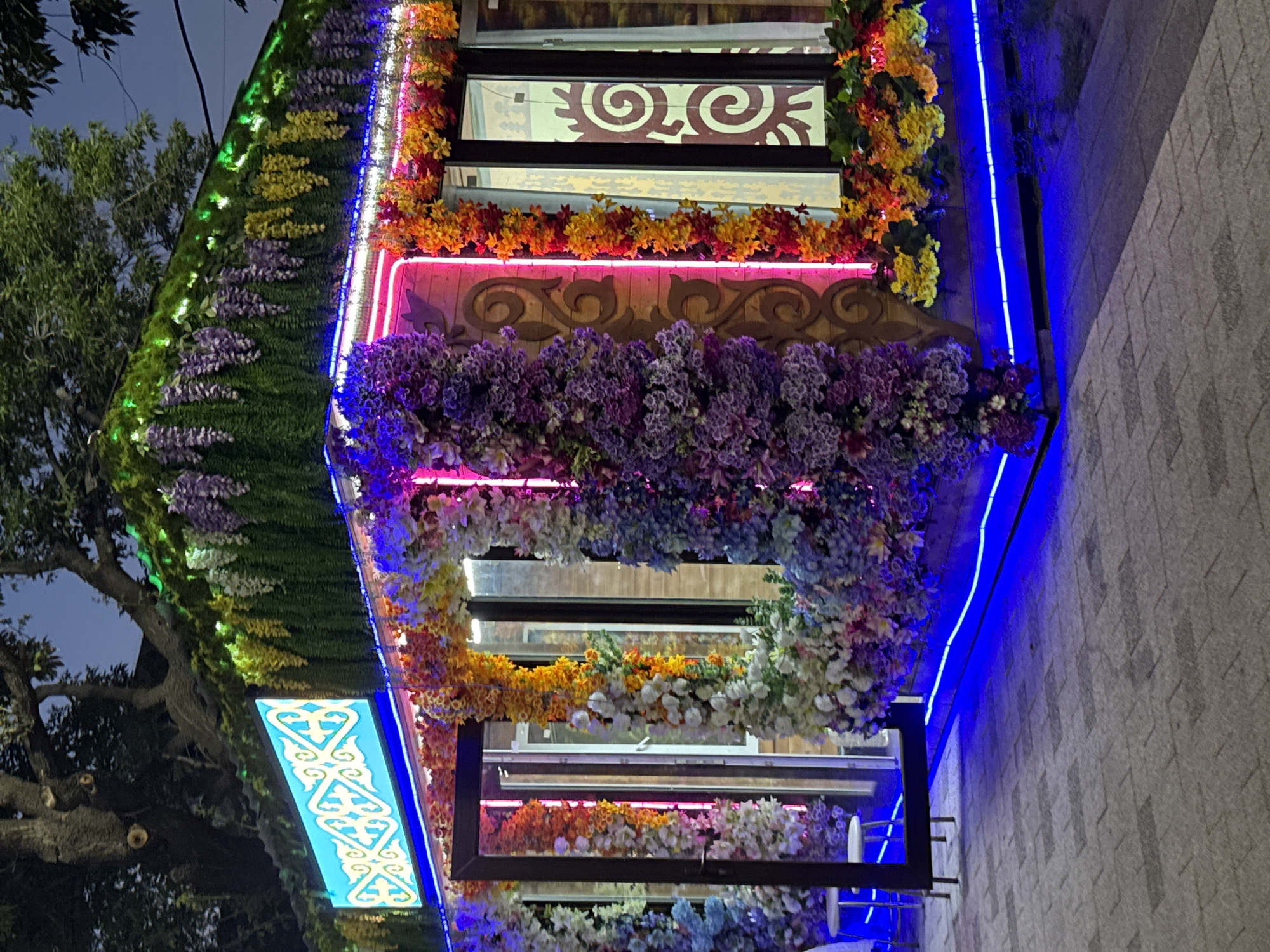 A brightly decorated kiosk or small building is illuminated by neon and LED lights at dusk. The structure is adorned with an abundance of artificial flowers in vibrant colors, including purple, blue, orange, yellow, and white, cascading down its sides and along its roofline. A large, illuminated blue sign with intricate white and gold patterns is mounted above the entrance. The building has glass doors and windows, with some revealing interior light. The ground in front is paved with grey interlocking bricks, and blue LED lights line the base of the structure. A large tree with dark branches is visible in the upper left portion of the image. The scene is located in the city of Petropavl, Kazakhstan. No people are visible in the image.