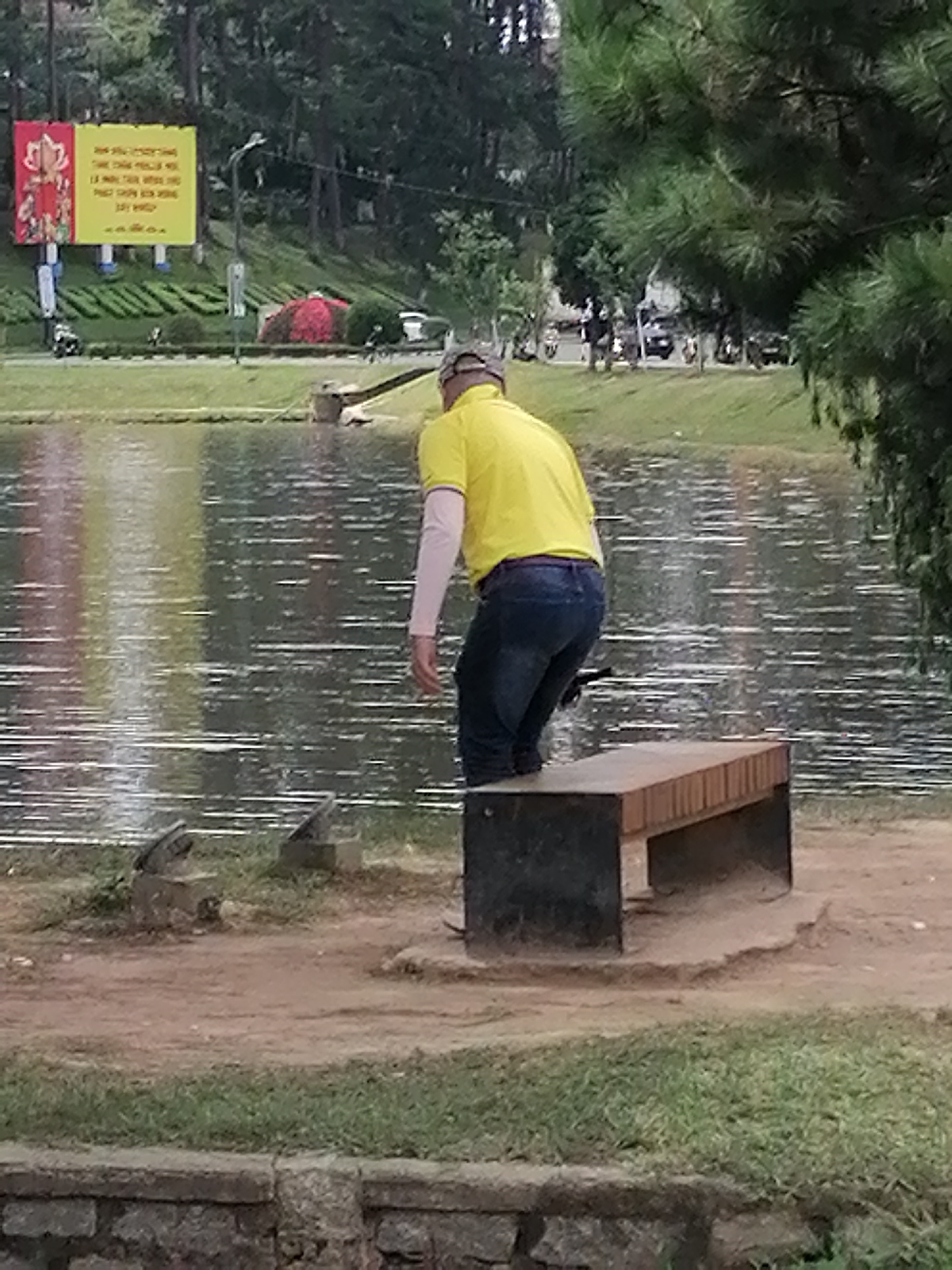 The image depicts a scene at Xuan Huong Lake in Da Lat, Vietnam. A person wearing a yellow t-shirt and blue jeans is standing by a wooden bench next to the lake. The person appears to be leaning forward, possibly interacting with something on the ground. The lake's surface reflects the surrounding trees and sky. In the background, there are more trees, a billboard with Vietnamese text, and some parked vehicles. The area around the bench is a dirt path with some grassy patches.