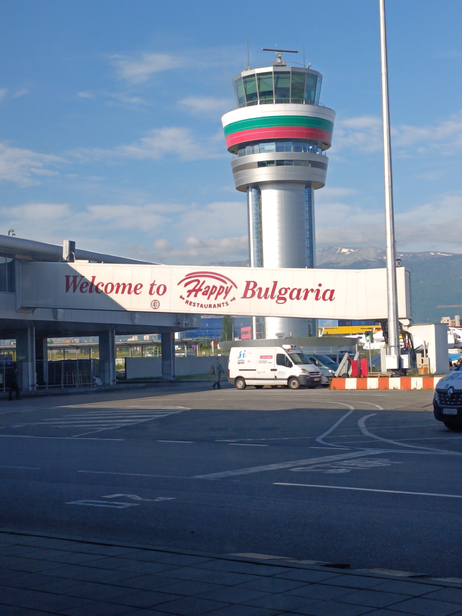 The image depicts an outdoor scene at Sofia Airport (SOF) in Sofia, Bulgaria. Dominating the background is the airport's air traffic control tower, painted with the colors of the Bulgarian flag.
In the foreground is an elevated pedestrian walkway with "Welcome to Happy Bulgaria Restaurants" displayed in red lettering. Below the walkway, a crosswalk and a road show directional arrows on the pavement. A white van is parked near a traffic barrier with red and white stripes. A person is walking along the walkway. A part of a dark car is visible on the right.
In the background, mountains are faintly visible. The sky is light blue with some clouds.