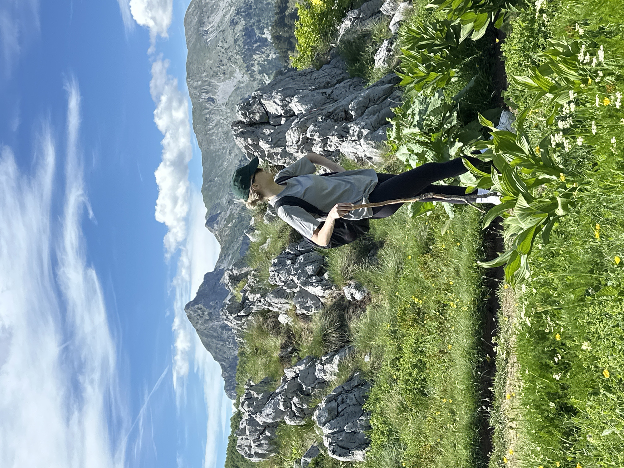 A young woman, wearing a green baseball cap, sunglasses, a grey t-shirt, black leggings, and white sneakers, stands on a grassy, rocky hillside overlooking a mountain range. She is holding a walking stick and has a black backpack on. The background features jagged grey rocks, green grass, and a bright blue sky with scattered white clouds. In the distance, imposing mountains with grey, rocky slopes rise under the sky. The overall scene depicts a person hiking in a natural, mountainous environment. The location is stated to be Dolja, Montenegro.