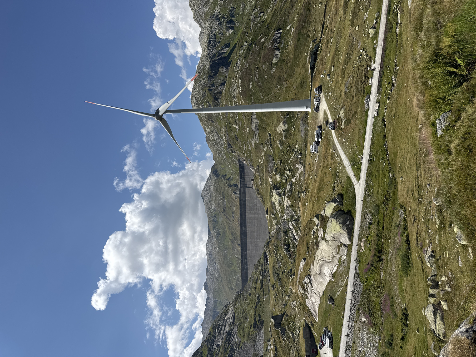 This image depicts a scenic view in the area of Airolo, Switzerland, likely at the Gotthard Pass, showcasing a blend of natural and industrial elements. In the foreground and midground, a vast, mountainous landscape unfolds, characterized by rocky terrain, green grassy slopes, and scattered boulders. A winding paved road traverses the scene, with several white recreational vehicles, possibly campervans, parked along its edges in the midground.
Dominating the right side of the image is a tall, white wind turbine, its blades angled upwards against a bright blue sky dotted with fluffy white clouds. The turbine stands as a modern addition to the rugged natural environment.
In the background, a massive concrete dam stretches horizontally across a valley, its imposing structure a testament to engineering. The surrounding mountains are rugged and appear sparsely vegetated, with rocky outcrops and shadowed crevices. A few small buildings, possibly associated with the dam or surrounding infrastructure, are visible near its base.
The overall impression is one of a high-altitude location where human infrastructure coexists with a grand natural landscape. The sunlight suggests it is daytime, and the clear sky indicates fair weather. There are no visible individuals engaged in significant actions, but a single person is seen walking along the road in the midground.