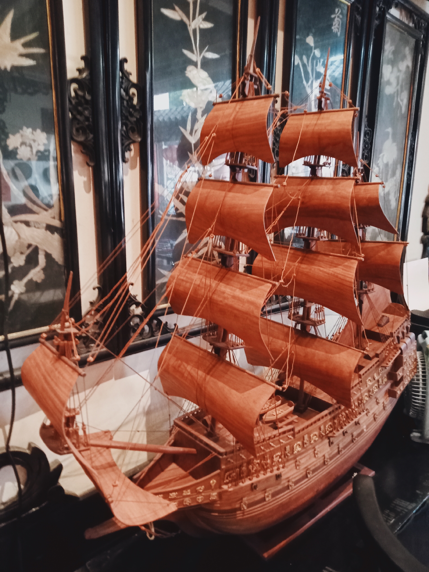The image displays a highly detailed wooden model of a multi-masted sailing ship, likely a historical galleon, positioned indoors. The ship model is predominantly reddish-brown, featuring numerous unfurled sails across its masts, intricately connected by a network of rigging. The hull exhibits fine carvings and decorative elements. The model rests on a wooden stand placed on a dark, flat surface, possibly a table or shelf.
Behind the ship, a series of tall, dark panels, resembling a traditional Asian folding screen or wall art, forms the background. These panels are embellished with light-colored motifs, possibly mother-of-pearl inlay or painted designs, depicting birds, branches, and flowers against a dark background, showcasing an elegant, traditional aesthetic. Dark, ornate metal fixtures are visible between some of the panel frames. Faint, calligraphic text or a symbol can be observed on one of the background panels, though it is illegible.
The scene is a static display, focusing on the craftsmanship of the ship model and the decorative elements of the background. The interior setting, with its traditional Asian decor, aligns with the context of Nha Trang, Vietnam. The lighting is even, illuminating the intricate details of the ship and the art pieces.