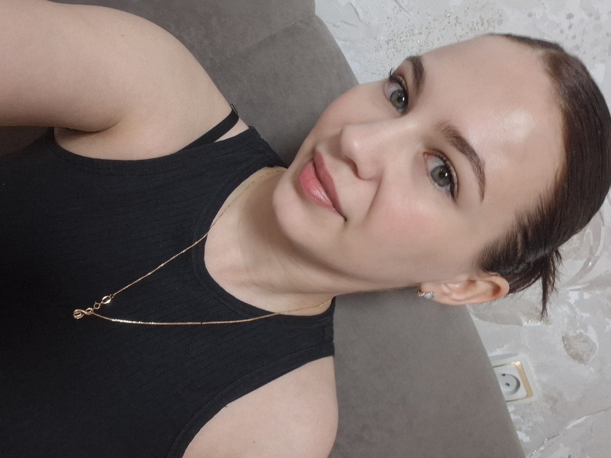 A woman is depicted in a close-up, angled shot, likely reclining on a grey upholstered surface such as a sofa or cushion. She has light skin, green eyes, and her dark hair is pulled back from her face. Her expression is gentle and serene, with a slight, pleasant smile. She is wearing a black ribbed tank top or bodysuit. Around her neck is a delicate gold chain necklace featuring a small, stylized infinity-shaped pendant. A small, light-colored hoop earring is visible in her right ear. The background shows a textured white wall, which appears to have some worn or peeling areas, and a white electrical wall socket with a gold-colored border. The setting is indoors, characteristic of a casual home environment in Chaikovsky, Russia. No visible text is present in the image.