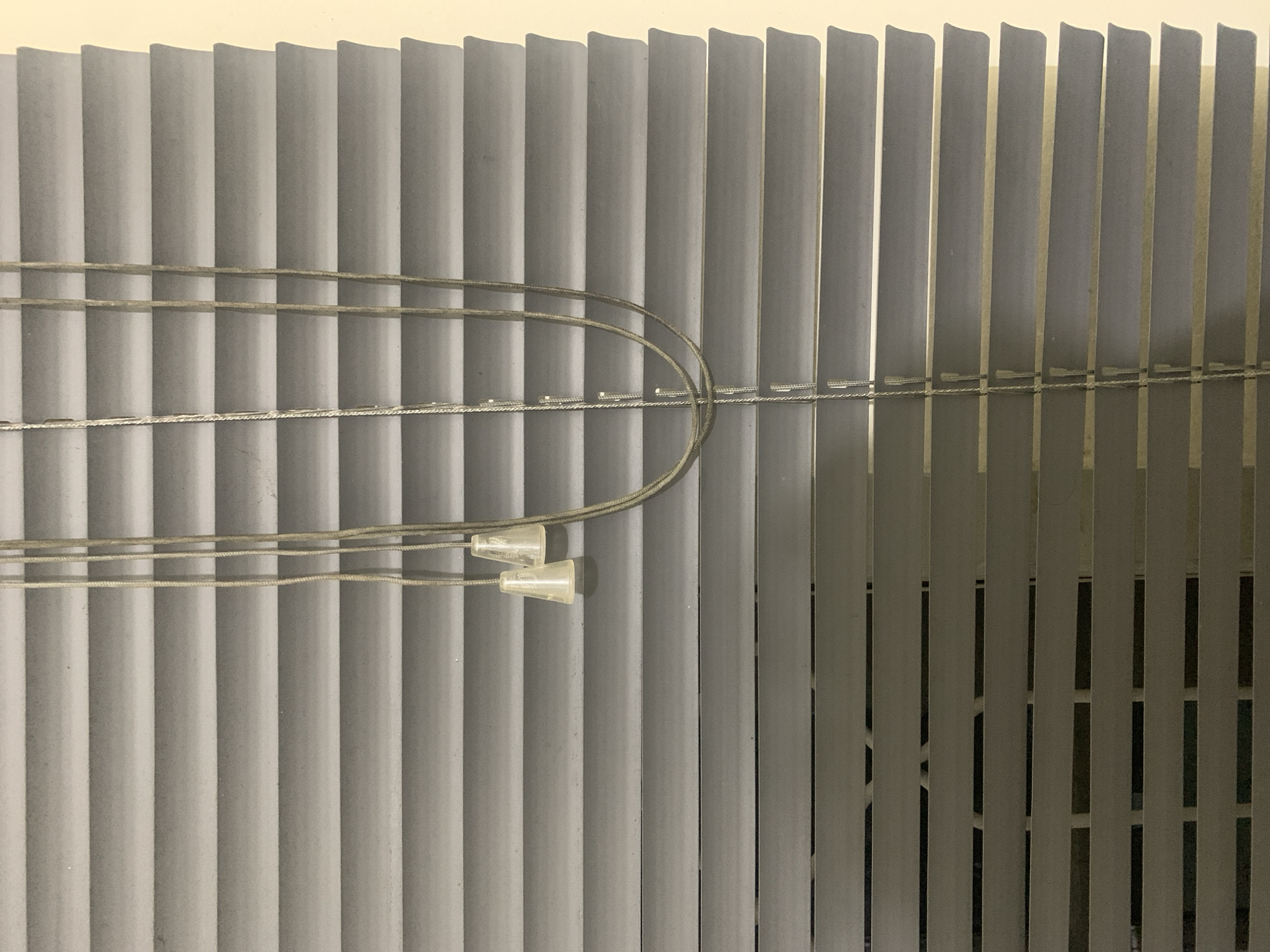 The image shows a close-up of vertical blinds. The blinds are a light gray color and appear to be made of plastic or a similar material. They are arranged in parallel, with slight gaps between them, suggesting they might be partially open or a type of louvered blind.
There are two thin cords running horizontally across the blinds. These cords are also gray and appear to be made of a braided material. Attached to these cords are two small, clear plastic cone-shaped objects. These are likely the pull cords or tassels for operating the blinds. The cords are looped around some of the blinds, indicating they are part of the blind's mechanism.
The background behind the blinds is not clearly visible, but it appears to be a wall or a surface that is a lighter shade than the blinds, possibly a light cream or beige color. The lighting is somewhat diffused, casting soft shadows on the blinds. The overall impression is of an indoor setting, possibly a room with windows covered by these blinds. There is no visible text in the image. The setting is indoors, and the time of day and weather are not discernible. The scene is static, with no apparent activity taking place other than the inherent function of the blinds.