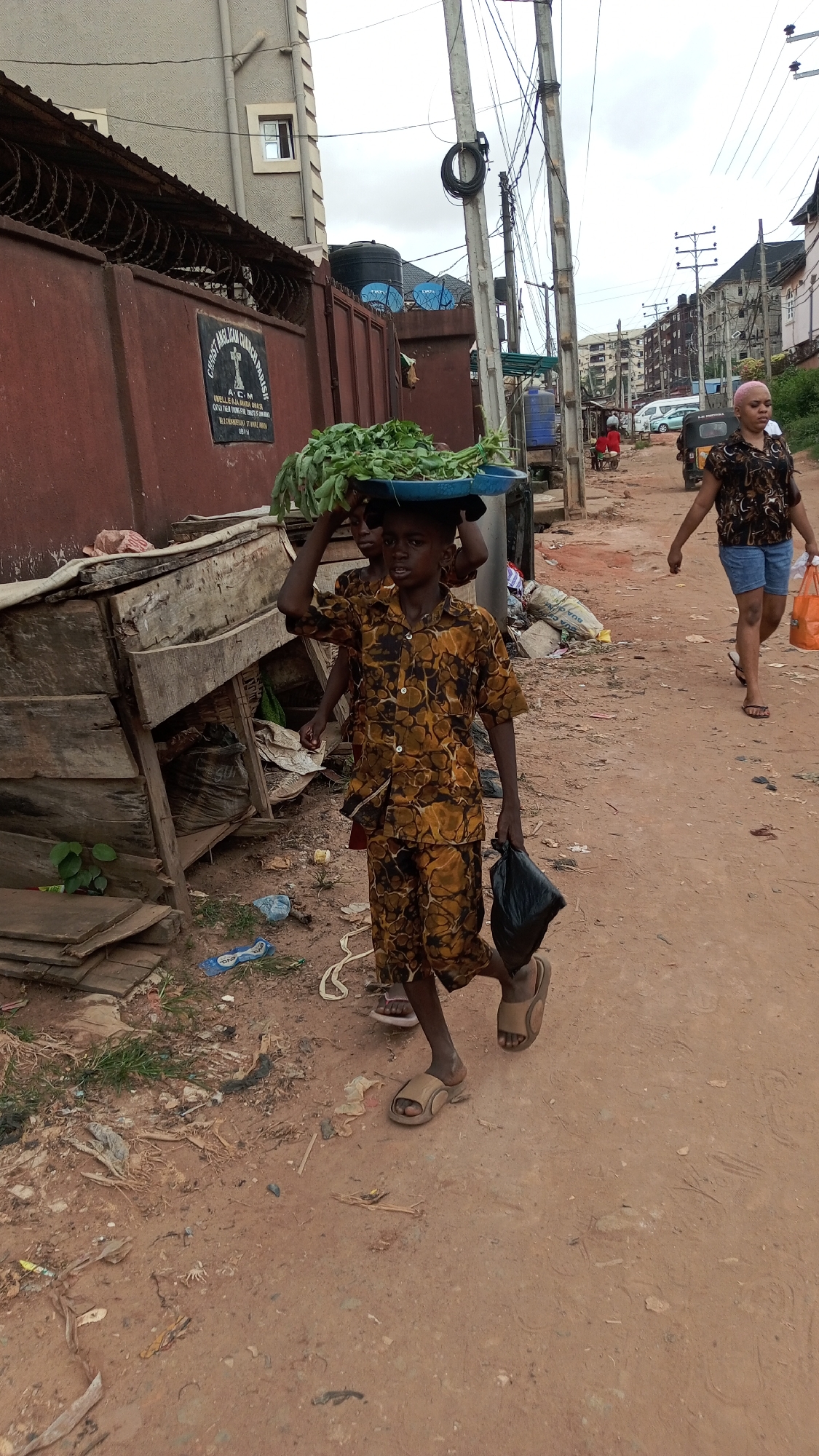 A young boy in a brown and yellow patterned shirt and shorts is walking on a dirt road in Obosi, Nigeria. He is carrying a tray of leafy green vegetables on his head and a black plastic bag in his hand. Another child is walking behind him, also appearing to carry the tray. A woman with a shaved pink head is walking in the opposite direction, carrying a shopping bag.
The road is littered with trash, and there are dilapidated wooden structures on the left side of the road. In the background, there are buildings, power lines, and parked cars. A sign on a nearby building reads "CHRIST ANGLICAN CHURCH, OBIELI AJAH OWA OBO, CRADLE YOUR YOUNG FOR CHRIST'S KINGDOM, ELDER NKEMDURIM EZE, VICAR, OBIELI OWA."