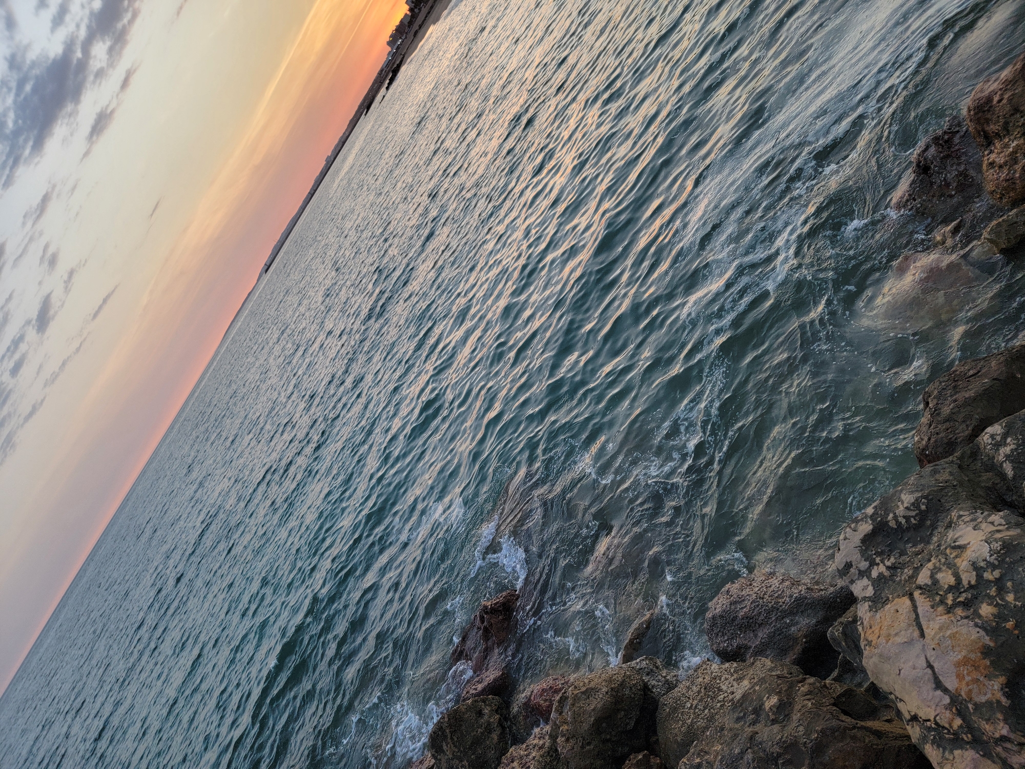 The image captures a seascape at dusk, with the horizon line angled diagonally across the frame. The foreground is occupied by rough, rocky shoreline, with large boulders in various shades of brown and grey. Gentle waves lap at the rocks, creating white foam and ripples on the water's surface. The water itself is a deep teal color, reflecting the fading light. The sky above transitions from a pale pink and orange near the horizon to a muted grey with scattered clouds towards the top of the image. In the distance, along the shoreline, there appear to be some buildings and structures, suggesting a coastal town or city. The overall scene evokes a sense of tranquility and the end of a day. Based on the provided context, this scene is located in Quarteira, Portugal.