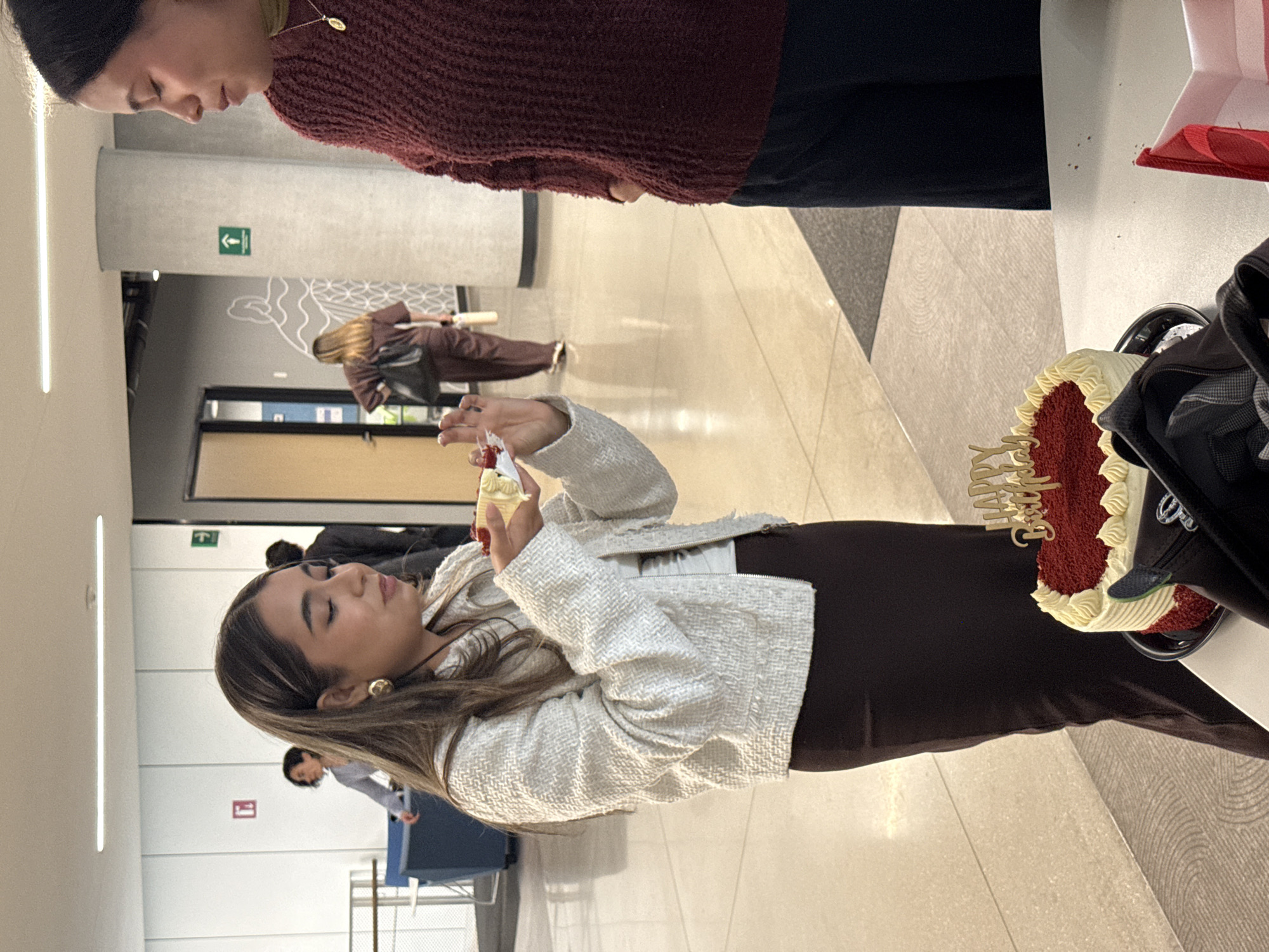 A well-lit interior space in Mexico City, Mexico, shows several individuals. One woman, in the center of the frame, wears a light-colored tweed jacket, a white top, and dark brown pants. She is holding a piece of cake topped with frosting and a red garnish. Next to her is a table with a red velvet cake with white frosting and a "Happy Birthday" cake topper. Another person is to the left of the woman in the light-colored jacket and is wearing a burgundy sweater with black pants. In the background, there are several people walking down the hallway, and there are visible exit signs. The scene suggests a birthday celebration or event in a public space, such as an office building or university hallway.