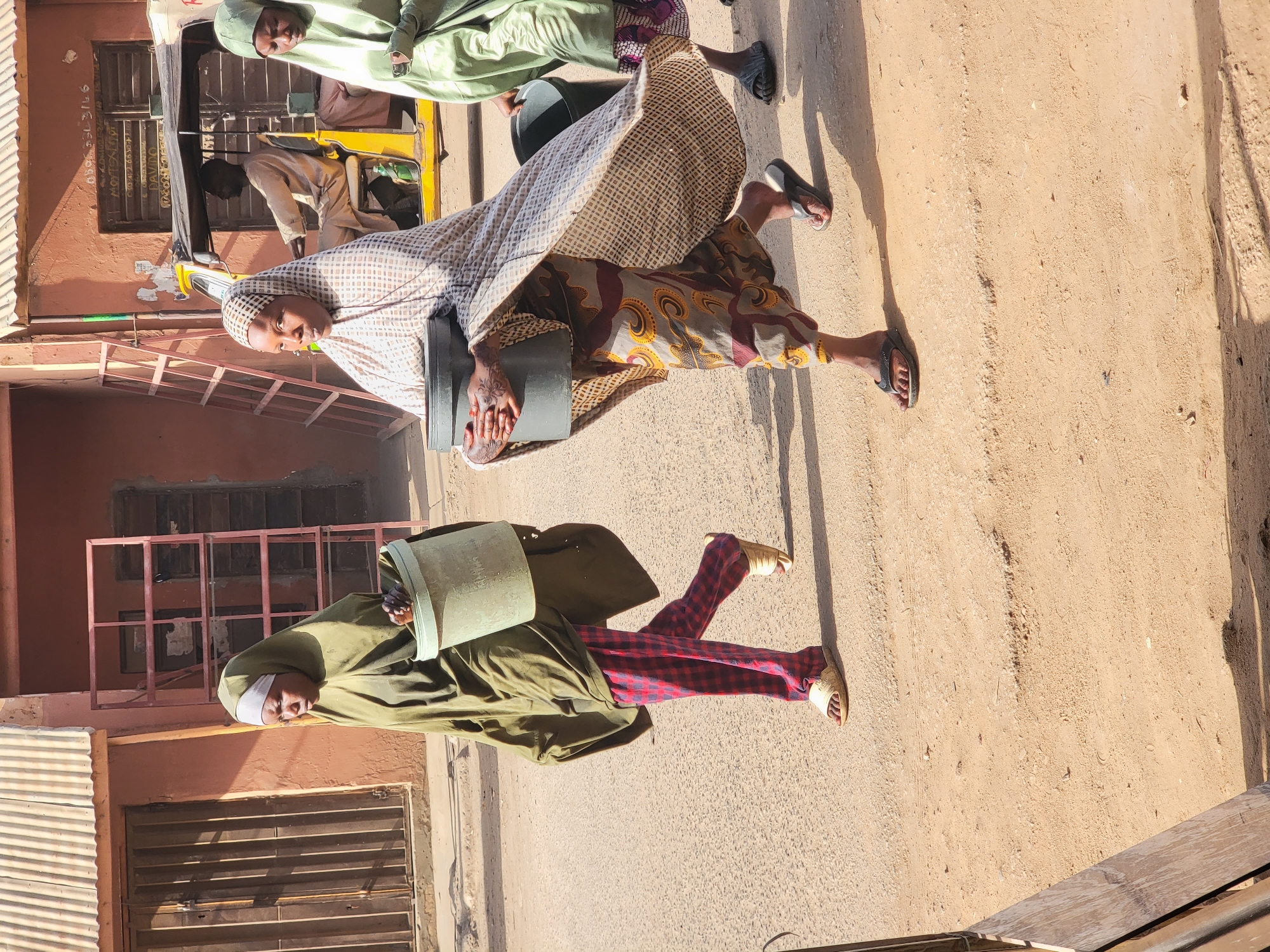 The image shows a street scene in Maiburiji, Nigeria. The ground is dirt. Two people are walking on the street. One person is wearing a long green robe and red and blue checkered pants and is carrying a green bucket on her head. The other person is wearing a long, patterned dress and pants and is carrying a black box under her arm. In the background, there is a parked yellow vehicle and a two story building with barred windows.