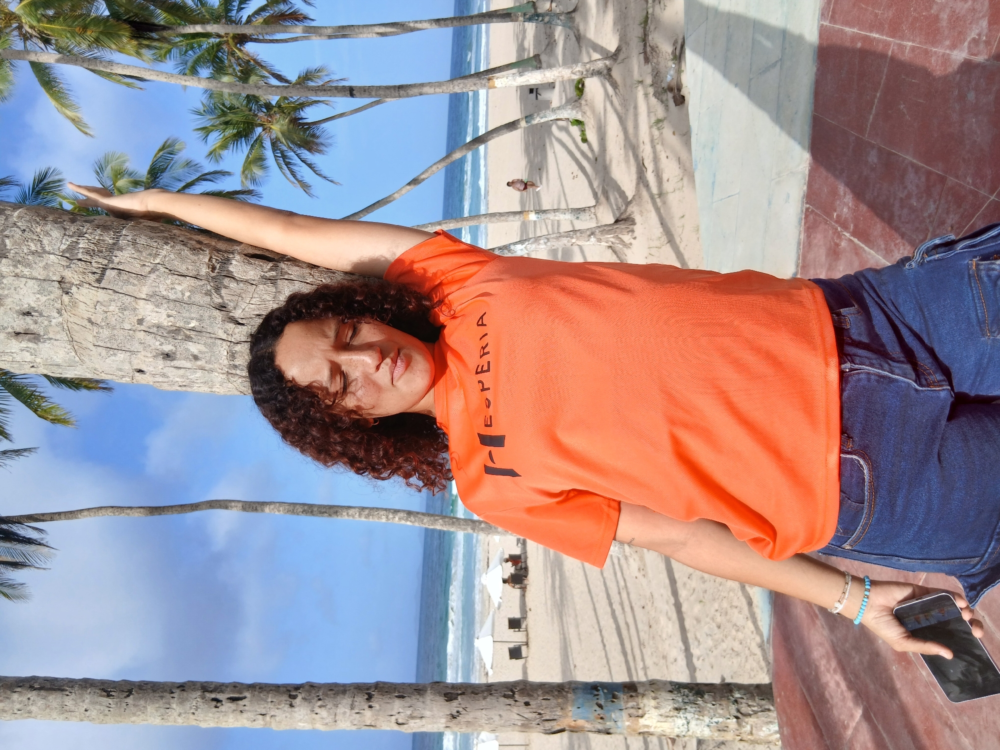 A young woman with curly brown hair is leaning against a palm tree on a beach, with her right arm raised and extended upwards. She is wearing an orange t-shirt with black text that reads "ESPERIA" and blue denim shorts. She holds a smartphone in her left hand, and a few bracelets are visible on her left wrist. The background shows a sandy beach with several palm trees, lounge chairs, and umbrellas. The ocean is visible in the distance, with small waves breaking on the shore. The sky is blue with some scattered clouds. The overall scene suggests a relaxed, sunny day at the beach, likely in a tropical location like Playa El Agua, Venezuela, known for its palm-lined shores.