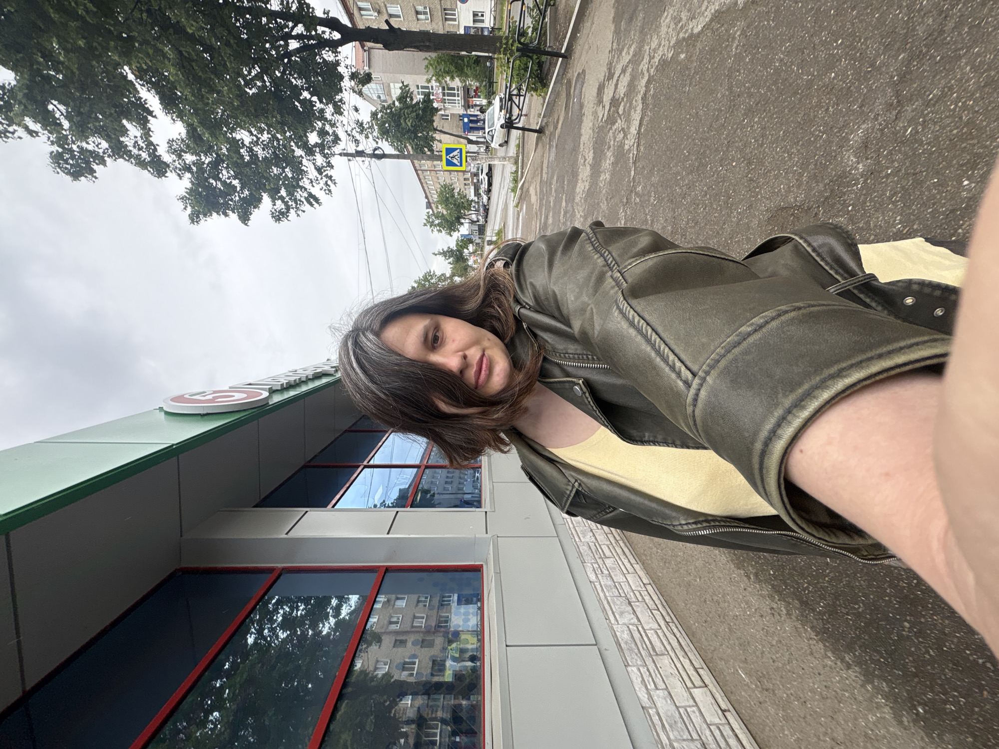 An outdoor selfie captures a person on a street in Chaikovsky, Russia, on an overcast day. The subject, a person with medium-length dark hair showing some grey strands, is seen from the chest up, wearing a dark olive-green leather jacket over a light yellow top. They have a neutral to slightly smiling expression, looking towards the camera.
In the background to the left, a modern building with a green roofline and several large windows with red frames is visible. The windows reflect the surrounding environment, including trees and a multi-story building. On the building's facade, a white sign with a red circular logo (resembling the Pyaterochka supermarket logo) and partial white Cyrillic text can be seen. To the right of the subject, an asphalt street or pavement stretches into the distance, lined by trees and a multi-story building. A yellow pedestrian crossing sign, mounted on a pole, is prominent in the mid-ground, indicating a zebra crossing. Parked cars are visible further down the street. The ground appears slightly damp in places. The overall setting is an urban environment, likely near a commercial establishment.