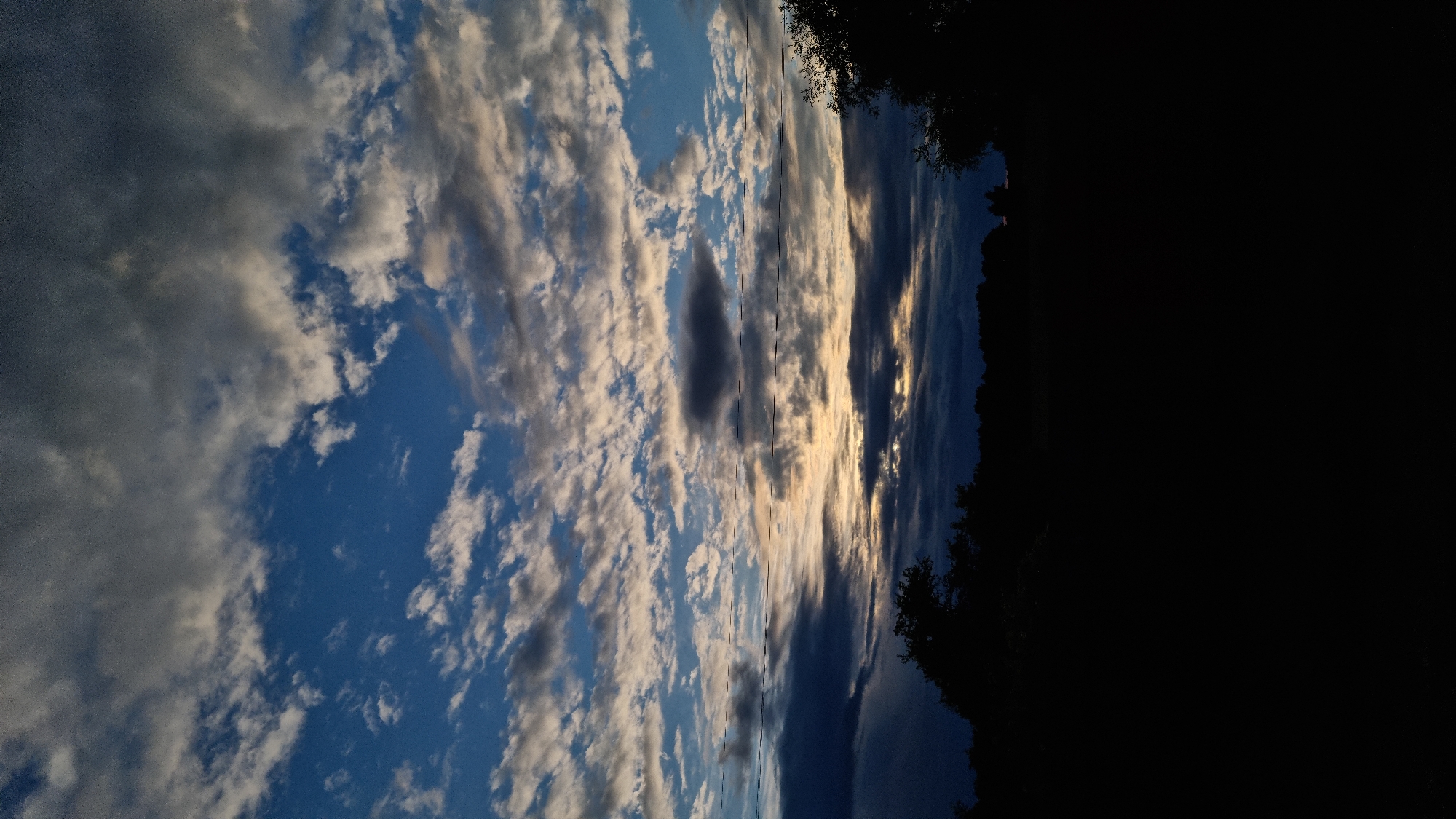 This is a vertical, full-frame photograph of a sunset over a silhouetted landscape. The sky is filled with a dramatic mix of dark, heavy clouds and lighter, wispy clouds interspersed with patches of bright blue sky. A horizontal band of warm, golden light emanates from the horizon where the sun is setting, illuminating the undersides of the clouds. In the foreground, the landscape is entirely black, obscuring any details of trees, vegetation, or terrain. There are no people or discernible objects in the scene. The overall impression is one of a natural twilight environment, possibly in a rural or park-like setting within Vysokoe, Russia, given the context.