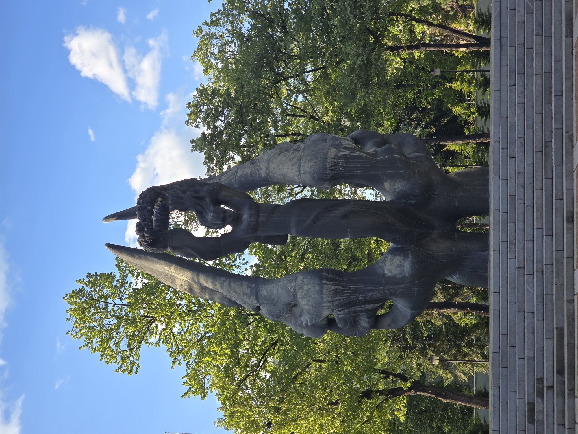 The image features a large, dark-colored, abstract sculpture set in an outdoor park or public space. The sculpture, possibly made of weathered metal such as bronze or steel, consists of several tall, intertwined organic forms that resemble stylized human figures or limbs. These elongated shapes exhibit a flowing, dynamic quality, reaching upwards towards the sky.
The setting is characterized by abundant green trees with lush foliage, suggesting a vibrant natural environment during spring or summer. Above the trees and the sculpture, a clear blue sky with scattered white, cumulus clouds is visible. The bright lighting indicates it is daytime, under sunny weather conditions.
To the right side of the sculpture, a structure composed of rectangular blocks of light grey stone or concrete creates a solid, architectural backdrop.
No people or activities are depicted in the scene, which focuses primarily on the sculpture's form and its natural surroundings. There is no visible text in the image.