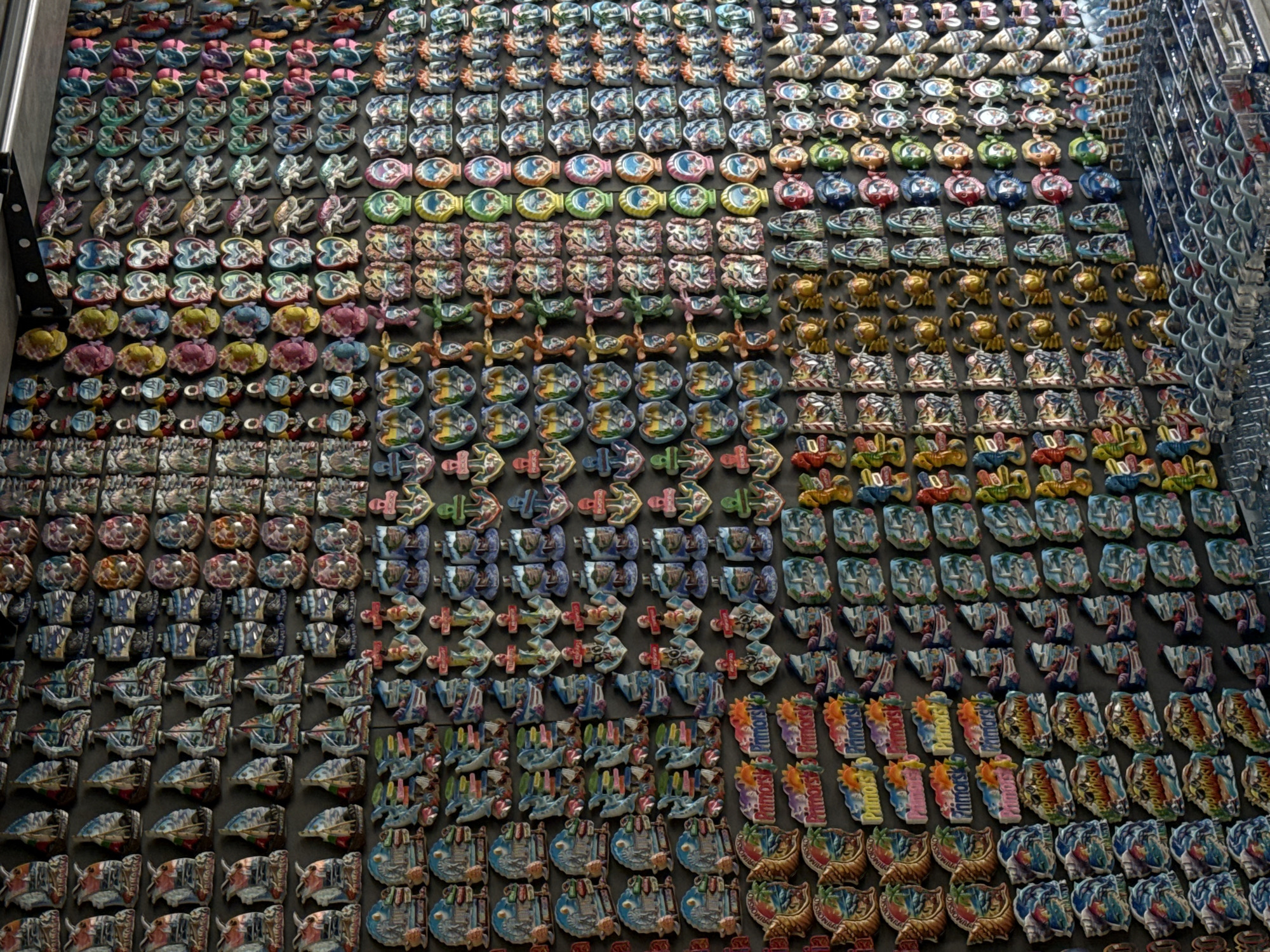 A vertical, eye-level shot captures a meticulously organized wall display of numerous souvenir magnets within a retail establishment in Primorsko, Bulgaria. The dark, uniform background highlights hundreds of individually affixed magnets arranged in precise horizontal and vertical rows. The top left portion of the display features multiple rows of stylized sailing ships in various colors. Directly beneath, rectangular magnets illustrate coastal landscapes, architectural scenes, and shell motifs, frequently inscribed with "Primorsko." Further down, the city name "Primorsko" is spelled out in large, colorful, individual letter-shaped magnets, interspersed with additional landscape and beach-themed designs, some containing the text "Primorsko Yarka."
The central and right sections of the display present a diverse array of magnet shapes and subjects. Prominently featured are multicolored anchors, golden crab figures, heart-shaped magnets with varied internal designs, stylized sea turtles, and small, cartoonish humanoid figures resembling gnomes or fishermen. A significant portion of the right side is dedicated to rows of circular and oval magnets exhibiting abstract patterns, colorful swirls, or simplified facial features. Toward the bottom right, vertical rows display brightly colored seahorse and stylized bicycle magnets. The lowest part of the frame reveals a multi-tiered transparent display system, likely constructed from acrylic or glass, indicating additional merchandise or presentation shelving below the primary wall display. No individuals are present in the scene.