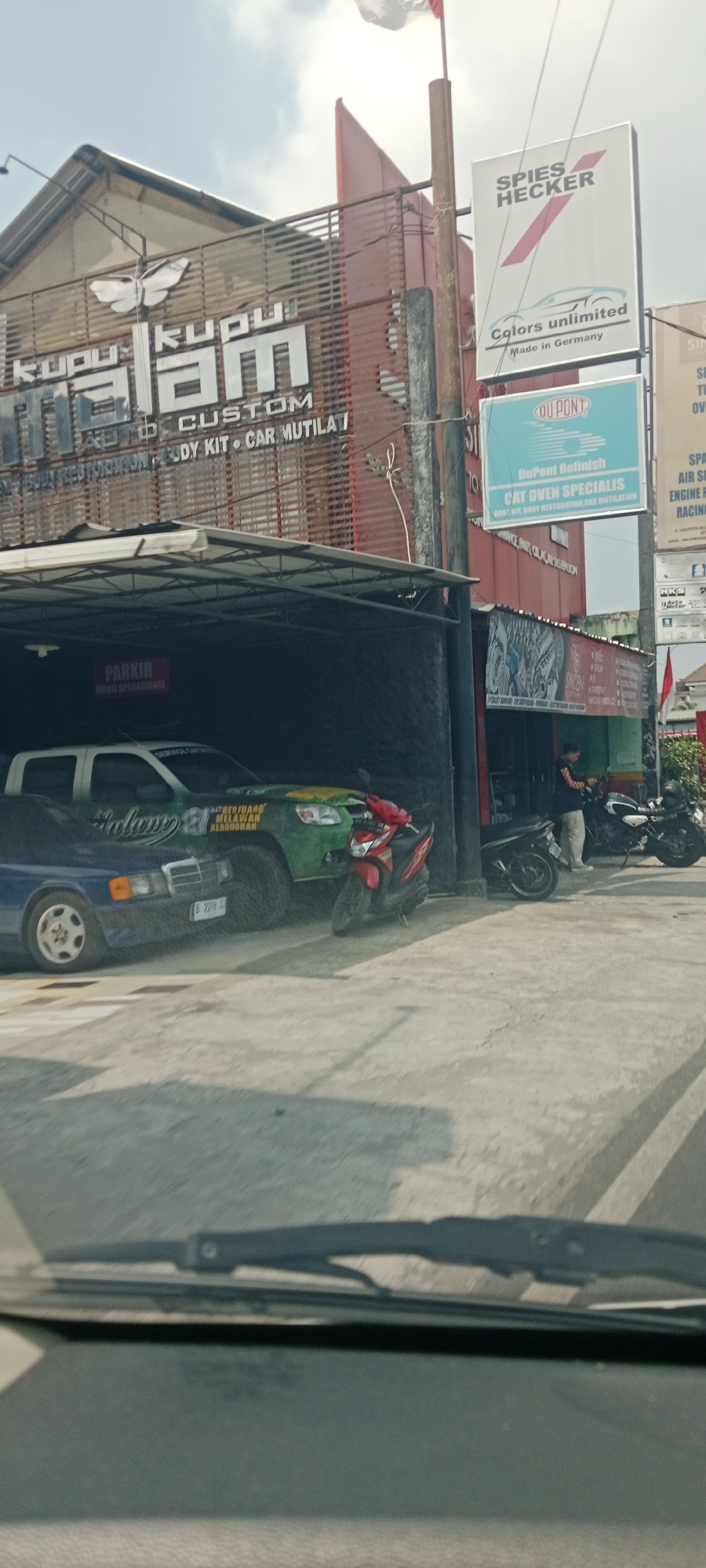 The image shows a car service and body shop located on Jalan Godean in Trihanggo, Indonesia. A large sign with the words "Kupu Kupu Malam" and "CUSTOM AUTO BODY KIT CAR MUTILATION" is visible on the building. Several vehicles, including a green pickup truck, a blue sedan, and two motorcycles, are parked in front of the shop. A man in a black jacket and light-colored pants is standing near one of the motorcycles. The sky is partly cloudy, and the sun is shining. The overall scene depicts a busy commercial area with a focus on automotive services.