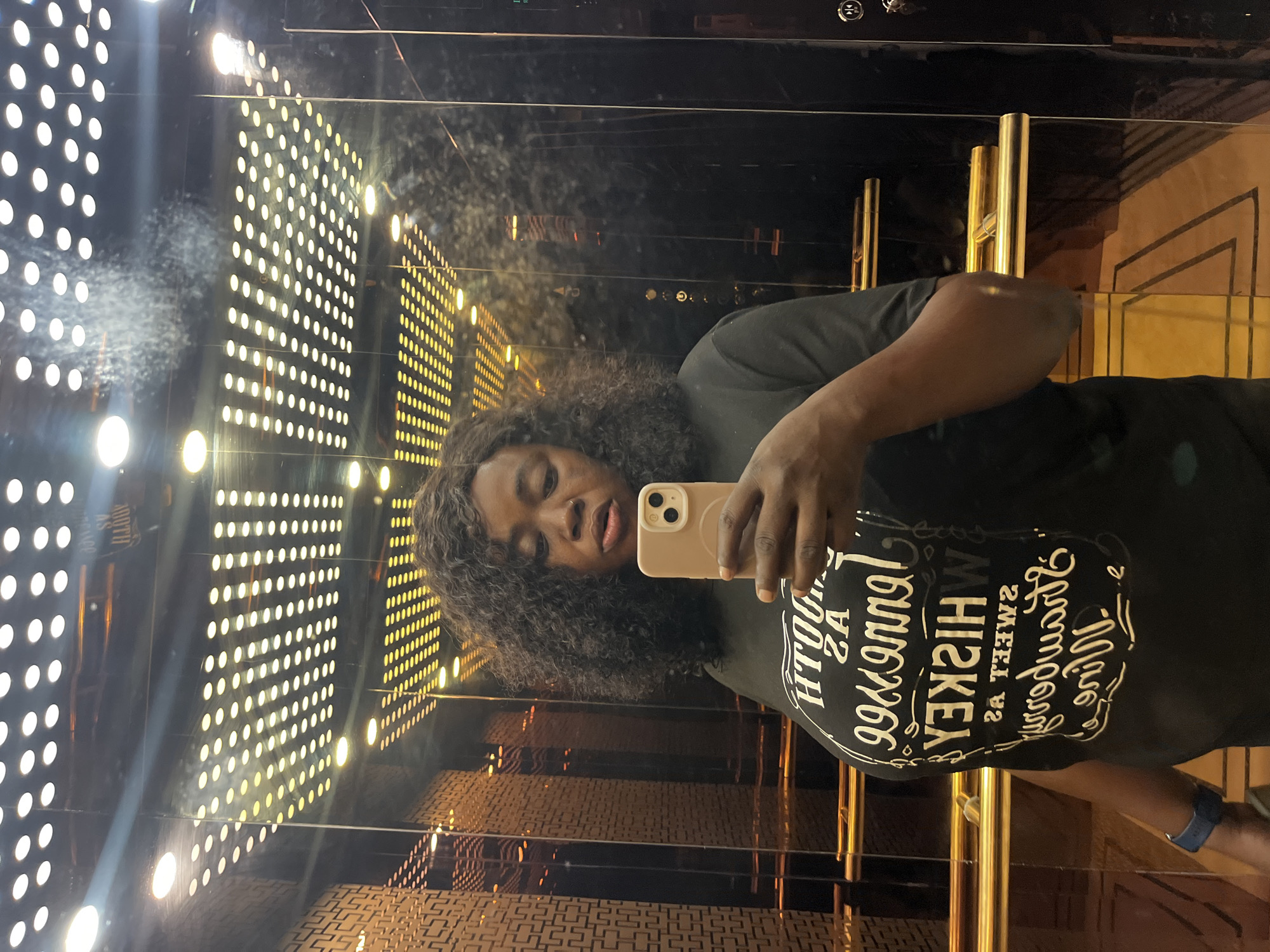A woman with dark, curly hair takes a mirror selfie in an elevator. She is wearing a black t-shirt with white text that reads "SOUTHERN AS TENNESSEE WHISKEY". She is holding a light pink smartphone in her right hand, capturing her reflection. The elevator walls are made of dark, reflective material with ornate gold handrails and decorative patterns on the side. The ceiling is illuminated by numerous small, circular lights. The floor appears to be tiled with a geometric pattern. The woman is standing on a patterned floor. The elevator is likely in a hotel or a modern building in Abuja, Nigeria, given the context.