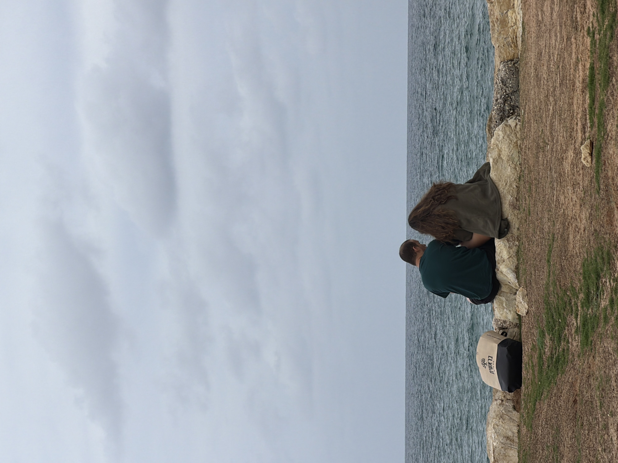 A couple is sitting on a rocky cliff overlooking the Mediterranean Sea in Tel Aviv, Israel. The woman has long brown hair and is wearing a green jacket. The man has short brown hair and is wearing a green t-shirt. They are sitting close together and looking out at the sea. A bag is placed beside them on the cliff. The sky is overcast.