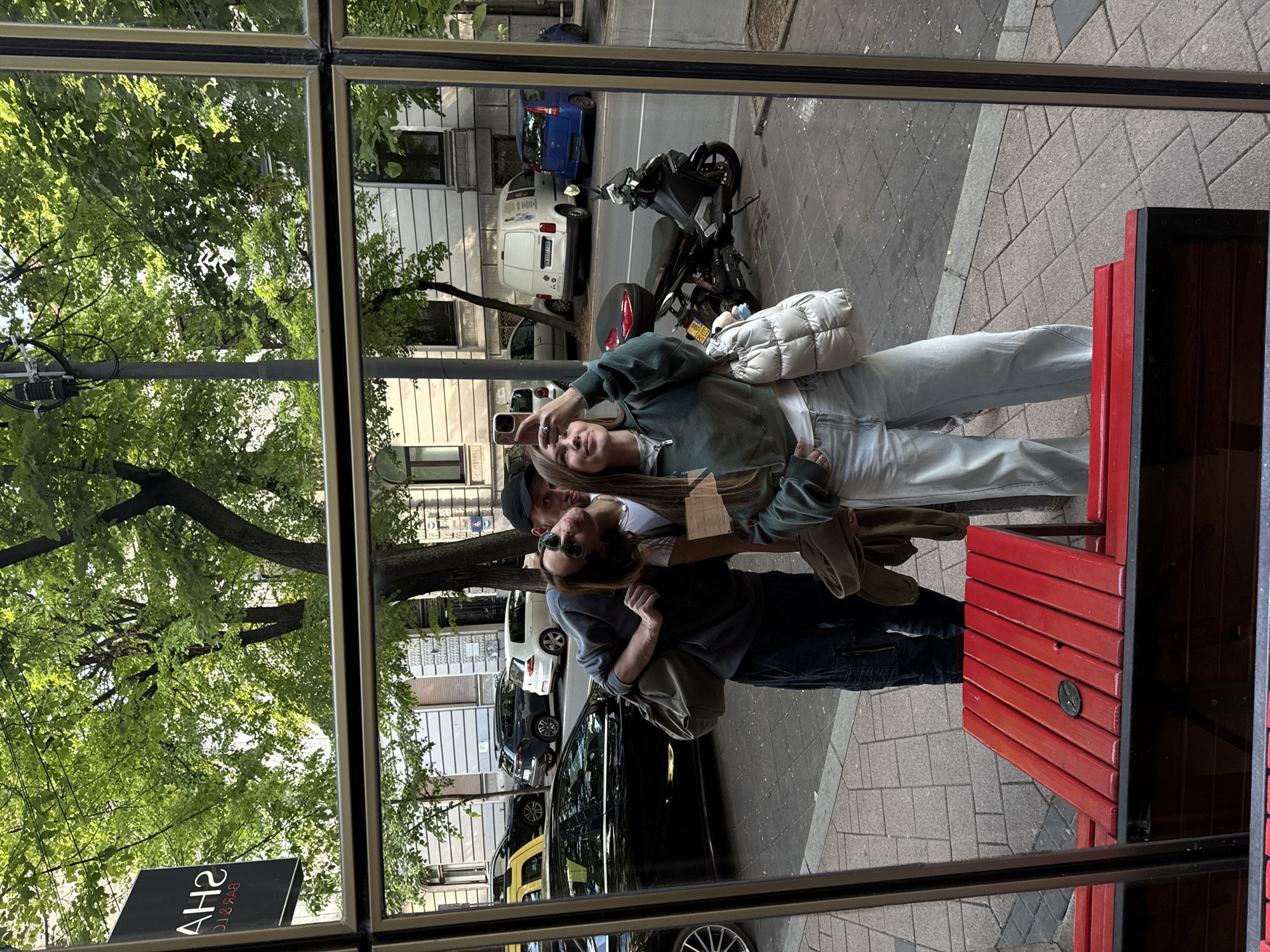The image captures a vibrant daytime urban street scene in Belgrade, Serbia, as a reflection in a window. The setting is characterized by a paved sidewalk, a street with parked vehicles, and multi-story buildings partially obscured by lush green trees, suggesting a pleasant spring or summer day.
Three individuals are the primary subjects, appearing to stand on the sidewalk just outside the window. In the center, a woman with long, light-colored hair, dressed in a dark green hoodie and light blue jeans, holds a smartphone up, seemingly taking a selfie or video. She carries a distinct white quilted shoulder bag. To her left, a man wearing a dark baseball cap and a white t-shirt stands with his arm around her. To her right, another man, wearing sunglasses and a grey long-sleeved top, also has his arm around her. All three individuals appear relaxed and content, with slight smiles.
Behind the group, a variety of vehicles are parked along the street, including a black scooter, a white van, a blue car, and a black sedan. The sidewalk features patterned grey and brown pavers. On the far right of the reflection, a red wooden structure, possibly a bench or planter, is visible. A utility pole with wiring extends vertically on the left.
Notable details include the bright natural light illuminating the scene, indicating daytime. A black sign, partially visible and mirrored in the bottom-left corner, clearly displays "SH2" and some smaller, mirrored text that resembles "BTK & BTC," likely part of a business name. The overall impression is one of a friendly, casual moment shared by the group in a lively city environment.