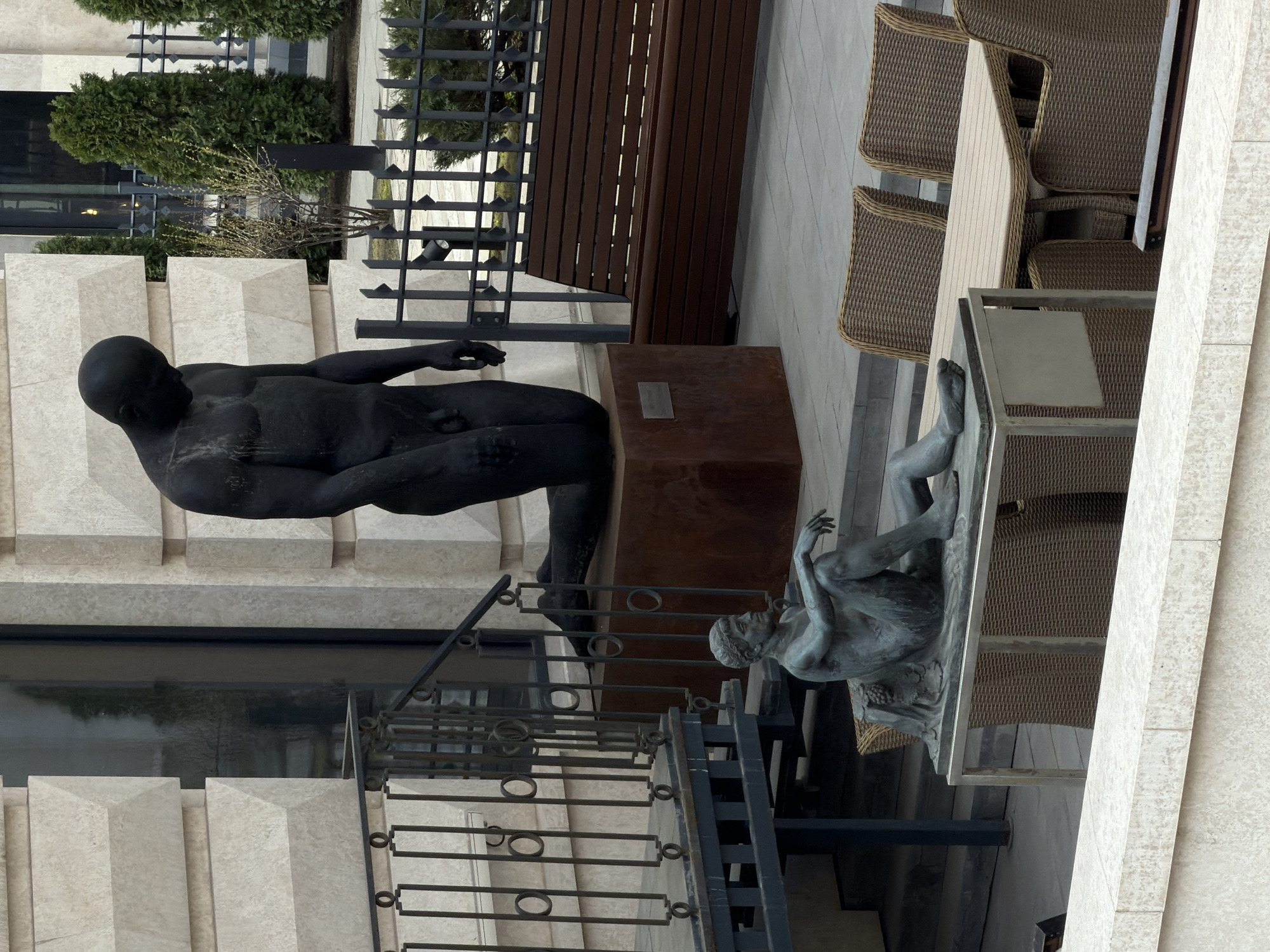 The image shows an outdoor scene in Moscow, Russia, featuring two sculptures in a public space. One sculpture is a large, black reclining male figure, positioned near a bench and a low barrier. The other sculpture, made of a lighter-colored metal, appears to depict a figure lying prone on a platform or table. Metal railings are positioned between the two sculptures. Wicker chairs and tables are nearby, suggesting an outdoor seating area. The architecture in the background includes stone walls with patterned relief. Green shrubbery is also visible.