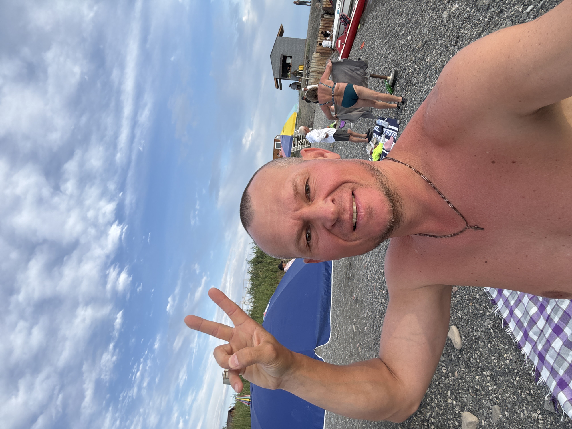 The image captures a selfie taken on a pebbly beach under a partly cloudy sky. In the foreground, a man with a shaved head, shirtless and showing signs of sunburn, smiles at the camera, holding up two fingers in a peace sign. He is wearing a silver chain with a cross pendant. Behind him, to the left, a large blue beach umbrella is set up. Further back, several people are visible. A woman in a blue bikini is seen from behind, holding a towel. Other individuals are standing or sitting near a small building with a grey facade and a light-colored roof. A red kayak and a white boat are also present on the beach. The scene suggests a relaxed day at the beach, possibly in the city of Zharbulak, Kazakhstan, given the reference.