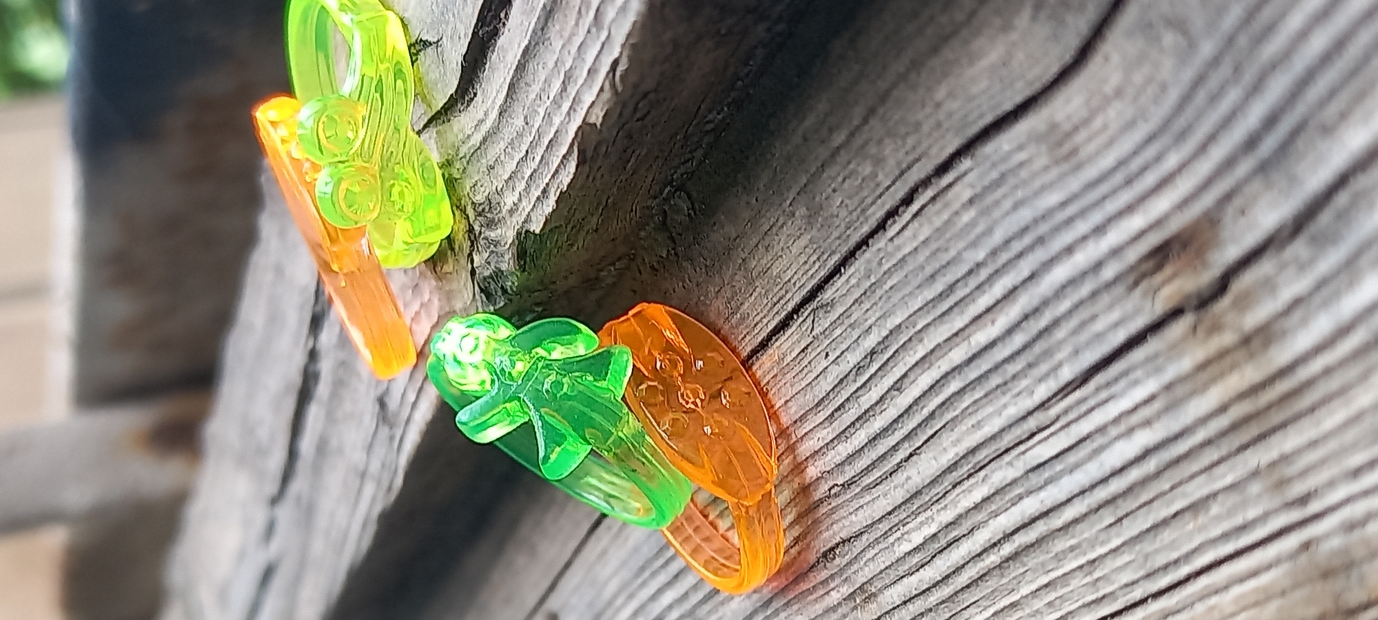 The image shows two brightly colored plastic toys on a weathered wooden surface. One toy, a green figurine with outstretched arms and a circular base, is positioned on top of an orange ring-shaped toy. The second toy, a bright green creature with multiple eyes, is partially visible behind and to the right of the first. An orange plastic piece, possibly part of another toy or a separate object, is situated above and to the right of the green creature. The wooden surface exhibits prominent grain patterns and appears aged. The background is blurred, suggesting a shallow depth of field. The image appears to be taken outdoors, with some greenery and a blurred structure visible in the distance. There is no indication of the city of Kryvyi Rih, Ukraine, in the image itself.