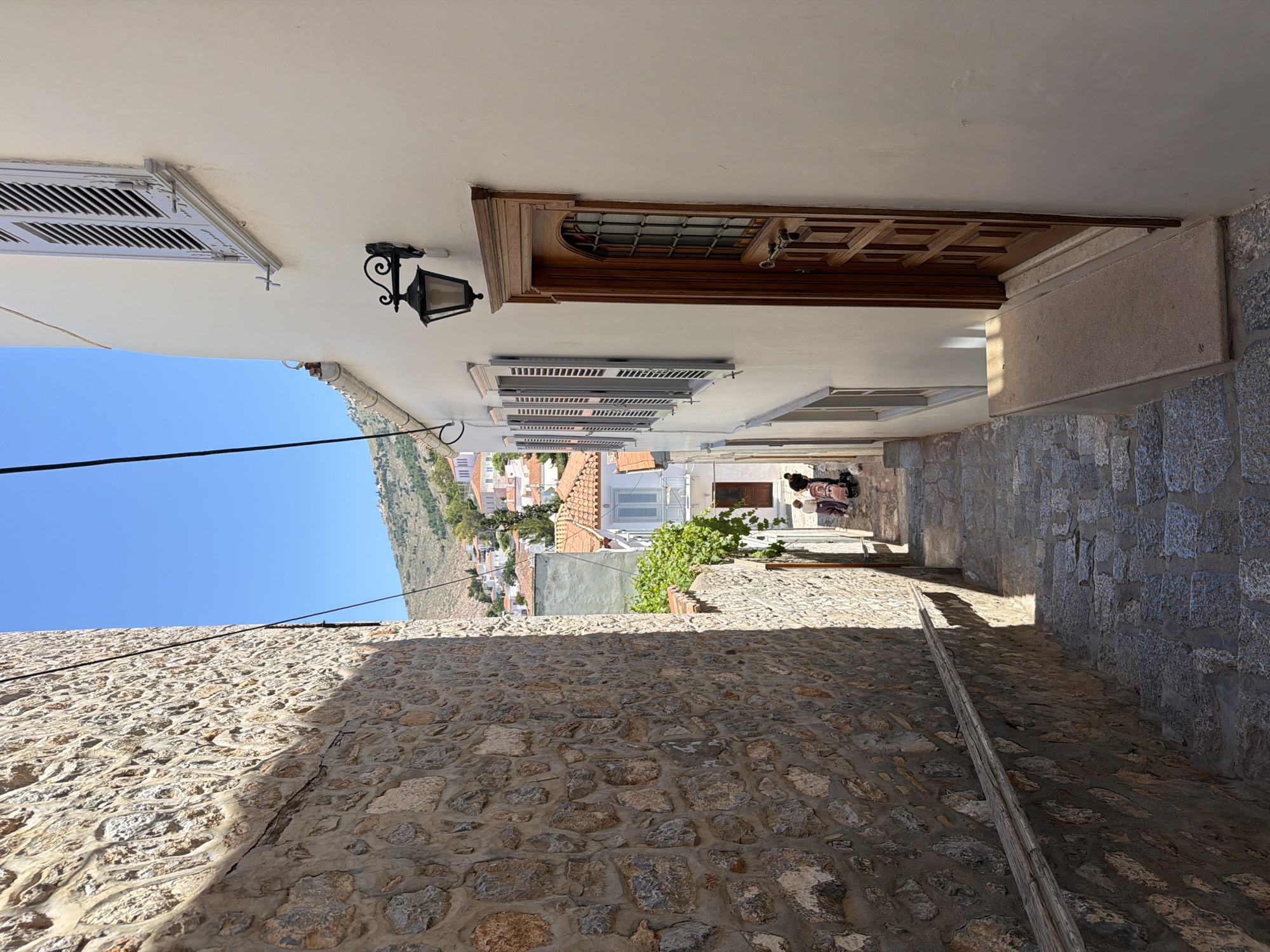 A narrow, shaded stone alleyway in Hydra, Greece, is flanked by a white building on the right with a wooden door and window shutters, and a tall stone wall on the left. The cobblestone street slopes downward into the light, where two figures stand with a stroller or luggage cart. In the distance, the town of Hydra extends up a hillside under a bright blue sky. A light fixture is mounted on the white wall. Overhead, a cable stretches across the alleyway.