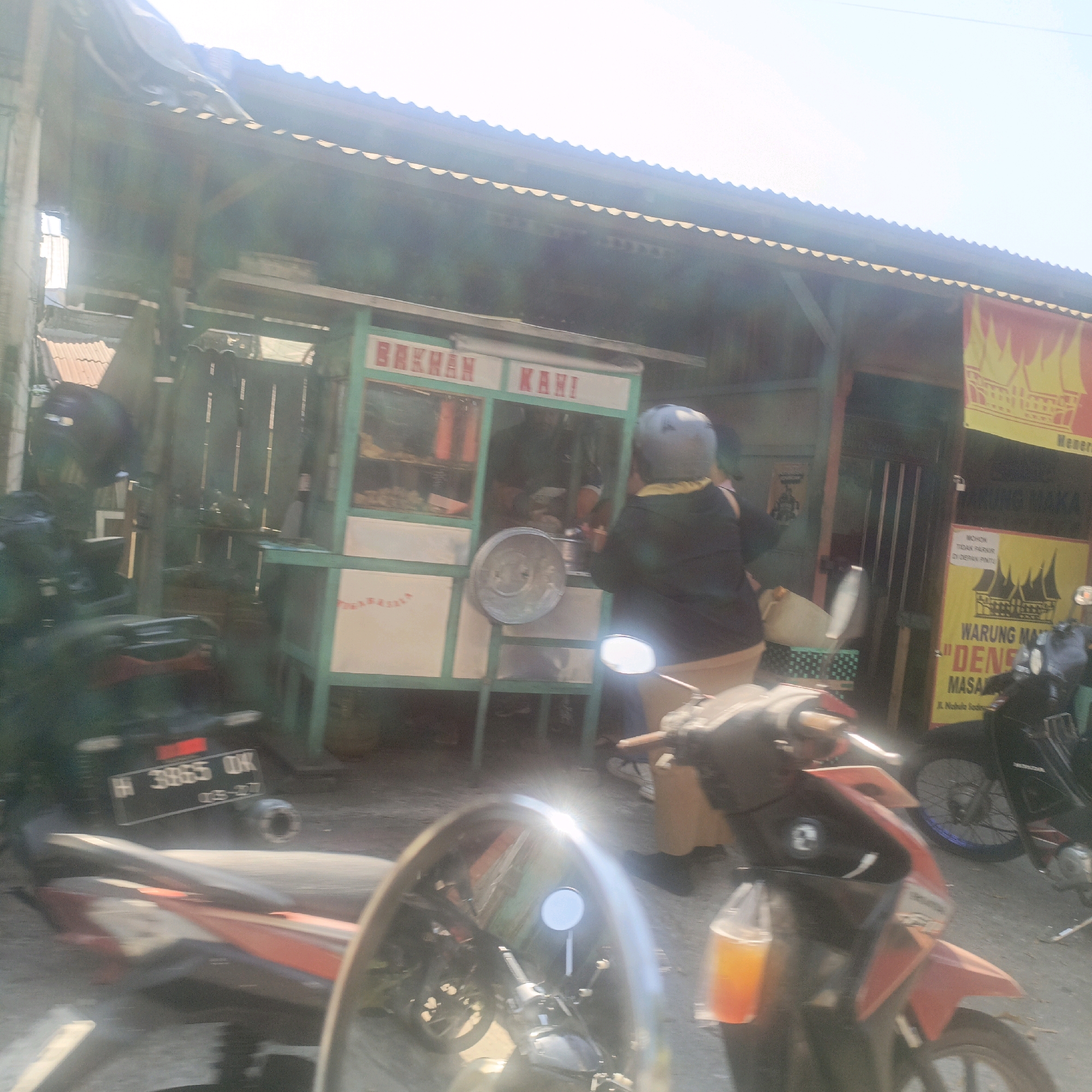 The image captures a street scene in Salatiga, Indonesia, likely in the daytime given the bright sunlight and clear sky. In the foreground, a red and black motorcycle is partially visible, its handlebars and a side mirror reflecting the surroundings. The motorcycle appears to be stationary or moving slowly. Behind the motorcycle, a food stall is set up, with the words "BAKMAN KAHI" visible on its facade. A person wearing a blue helmet and a dark jacket stands near the stall, seemingly interacting with the vendor. The stall is green and white, with glass panes displaying items within. To the right of the food stall, a yellow banner with red lettering hangs, advertising "WARUNG MAKAN 'DENS' MASAK". Another motorcycle, predominantly black, is parked behind the food stall, its rear wheel clearly visible. The background shows the facade of a building with a corrugated metal roof. License plate details visible on the motorcycle in the foreground include "H 3965 DK" and "02-277". The overall atmosphere suggests a typical day at a roadside food establishment in a local Indonesian setting.