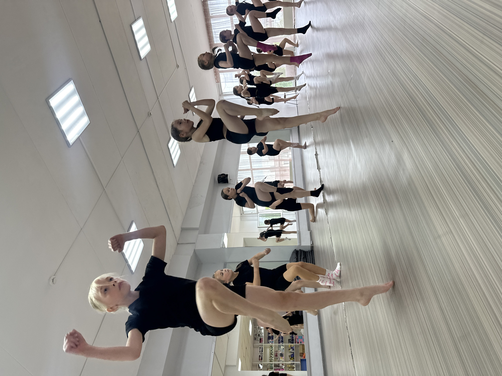 In a dance studio with a polished wooden floor and large mirrors, a group of young girls in black dance attire practice a routine. In the foreground, a blonde girl with her fists raised is mid-jump, her right leg bent at the knee and her left leg extended behind her. To her right, other girls are also in motion, some with legs raised in a similar pose, others in a lower stance. The room is lit by rectangular fluorescent ceiling lights. The studio appears to be located in Votkinsk, Russia, based on the provided context, though no specific Votkinsk landmarks are visible in the image itself. The overall impression is one of a focused dance class or rehearsal.
