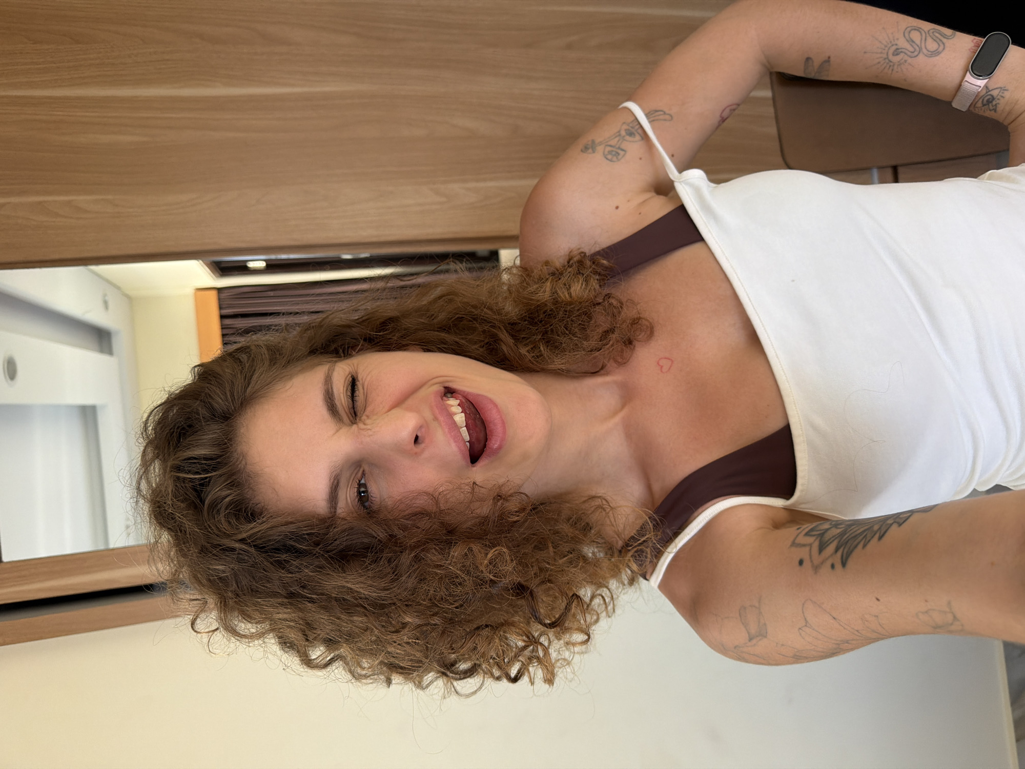 A young woman with curly brown hair, wearing a white tank top with brown straps, smiles and winks at the camera. Tattoos are visible on her arms. The background includes a wooden surface, a white wall, and a glimpse of a room with a door. The image appears to be taken indoors in North Kuta, Indonesia.
