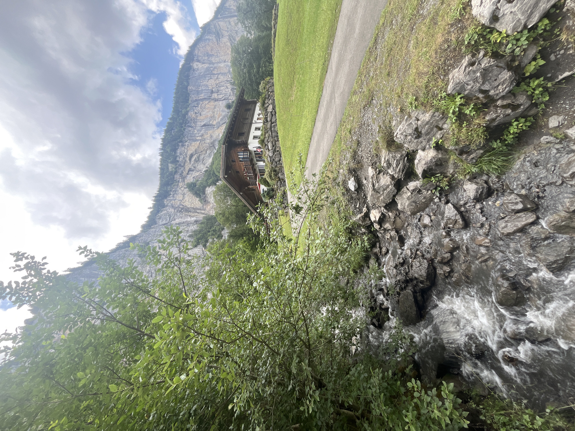 A wooden house with a red umbrella on a patio sits on a grassy hillside in Lauterbrunnen, Switzerland. A paved path winds up the hill toward the house. In the foreground, a stream flows over rocks, bordered by lush green foliage. The imposing Swiss Alps rise dramatically in the background, with a sheer cliff face and a lush green forest atop it. The sky is partially cloudy.