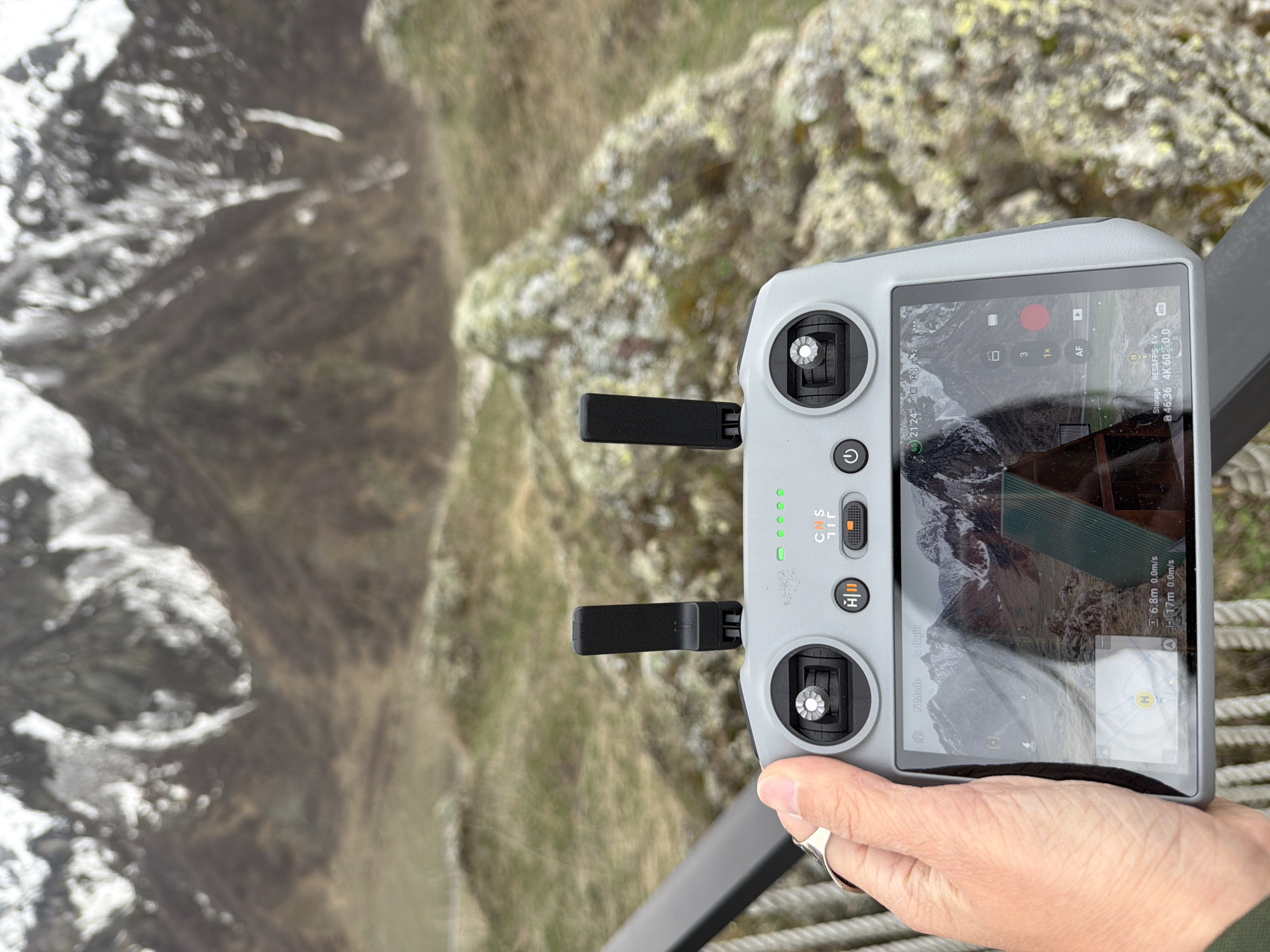 Here is a detailed description of the image:
**Scene:**
The image presents a close-up view of a drone controller held by a person against a backdrop of a mountainous landscape, likely in Pkhelshe, Georgia. The drone controller dominates the foreground. The person is holding the controller in their right hand.
**Drone Controller:**
The controller is gray and features various buttons, control sticks, and a screen displaying the drone's perspective of the surrounding environment. The screen shows a view of a mountainous area. The controller has 4 green lights on.
**Background Landscape:**
The background reveals a rugged mountainous landscape. There are patches of greenery mixed with rocky terrain and snow-capped peaks.
**Additional Details:**
The person is wearing a ring on their finger.
