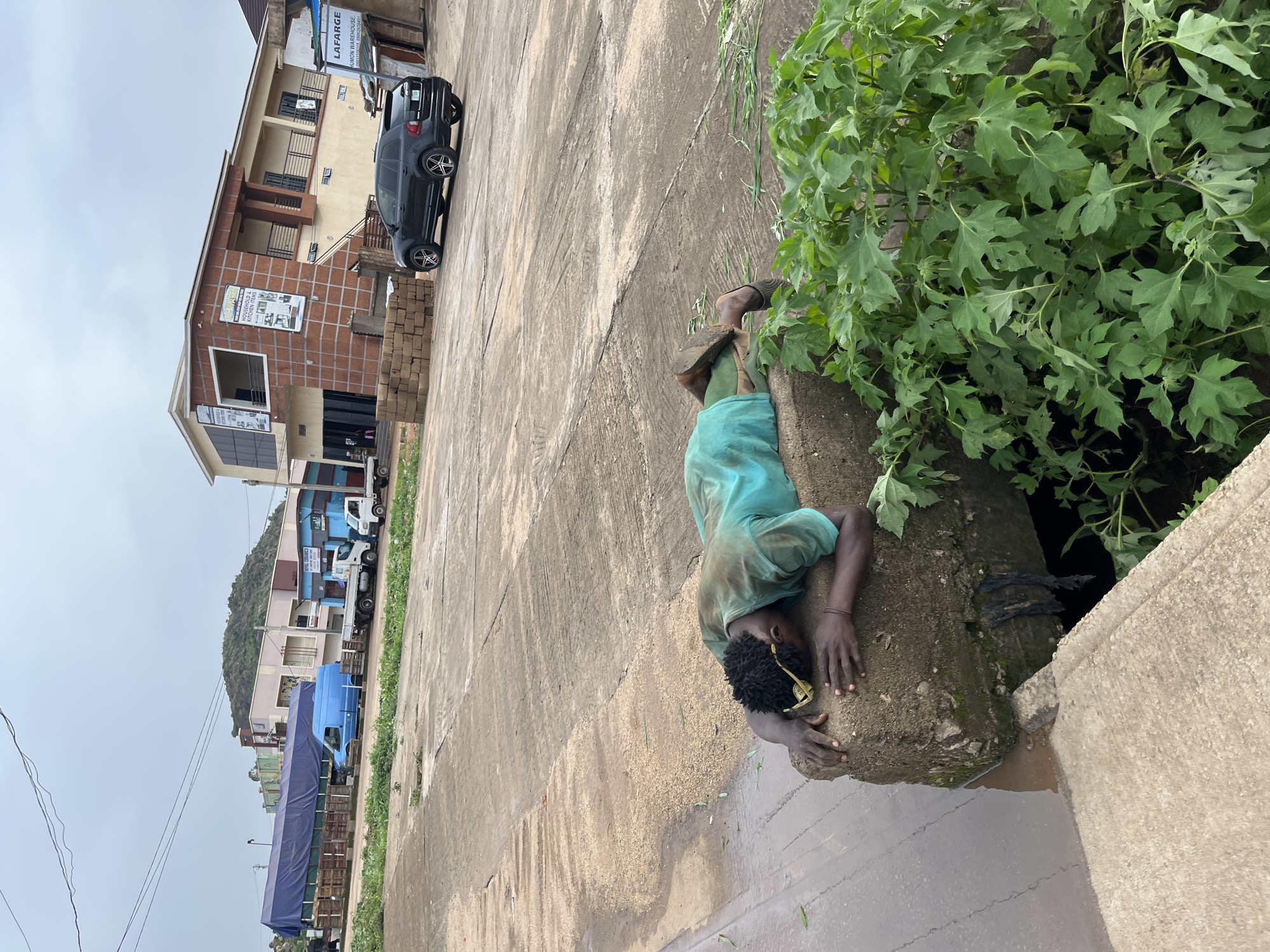 A person wearing a teal shirt lies prone on a concrete slab, their head resting on their hands. The person's face is obscured. They appear to be wearing light-colored sunglasses pushed up onto their forehead. The person's lower legs and feet are visible, wearing dark shoes. The concrete slab is positioned next to a drain with murky water. Lush green foliage is growing to the right of the slab. In the background, a dark-colored SUV is parked on a paved surface. Further in the background, several buildings are visible, including one with a sign that reads "LAFARGE." A blue tarp covers a structure to the left, with several trucks parked nearby. The sky is overcast. The location is identified as Gura Top, Nigeria.