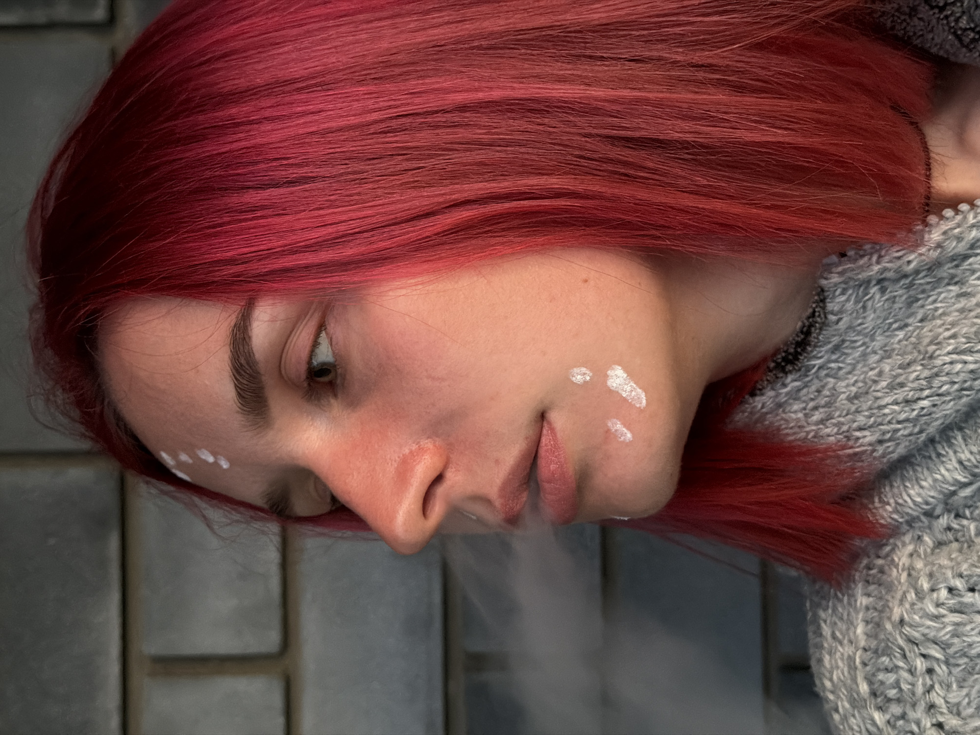 A close-up, profile view captures a young person with vibrant red hair against a blurred background of gray brickwork. The person's face, turned to the left, features a reddish flush across the cheeks and nose, with several small, irregular white markings on the forehead and cheek. The individual is exhaling a plume of white vapor from their lips. They are wearing a thick, gray knit sweater. The scene appears to be outdoors, with the brickwork suggesting an urban or semi-urban setting. The image does not contain any specific geographical markers that would allow for precise location within Chaikovsky, Russia.