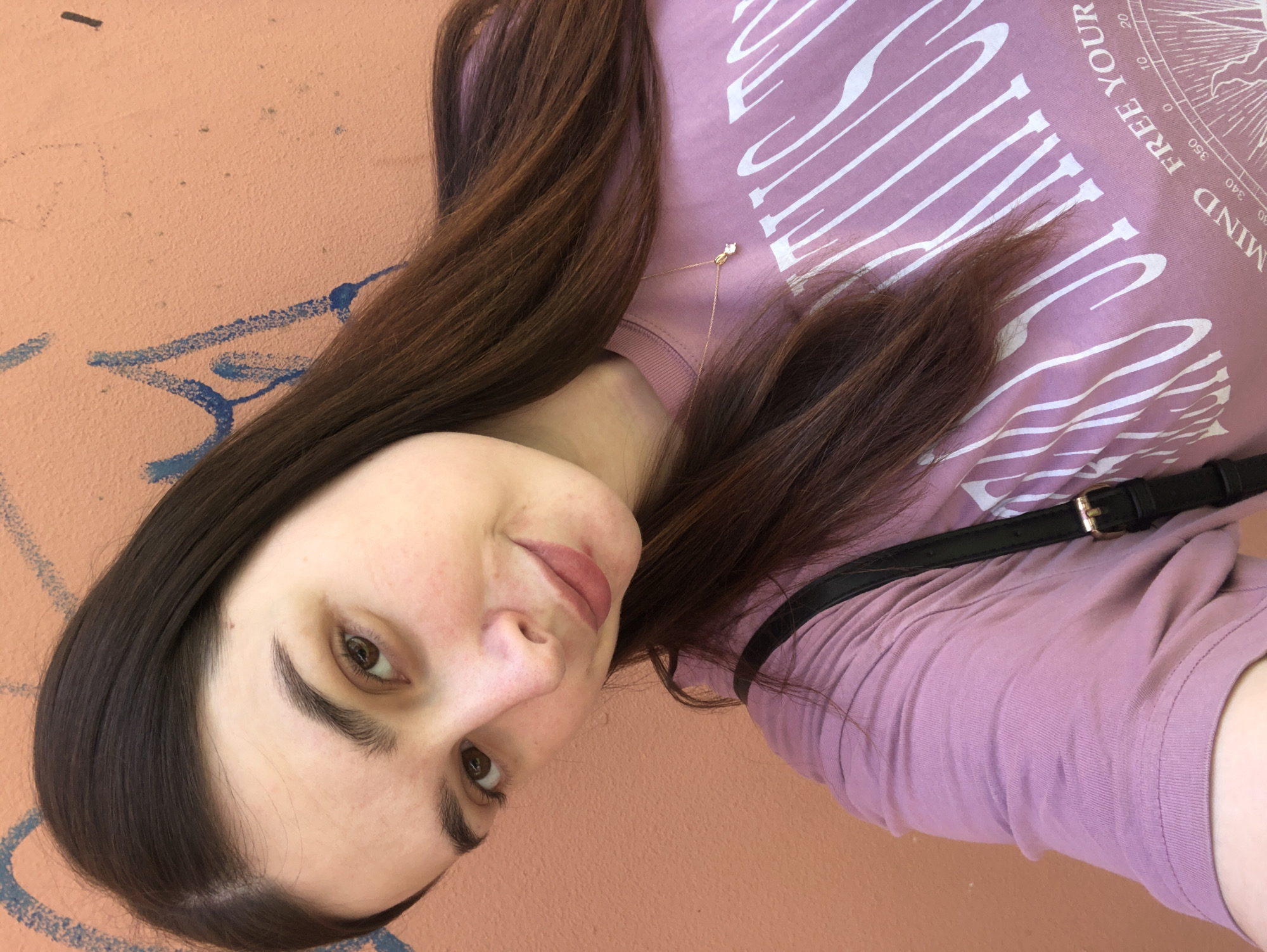 A young woman with brown hair is posing for a selfie against an orange wall with blue graffiti. She is wearing a light purple t-shirt with white lettering and a compass graphic. A thin gold necklace with a pendant is visible around her neck. A black purse strap crosses her body. The photograph appears to have been taken outdoors in Chaikovsky, Russia, as the information was provided within the context of that city.