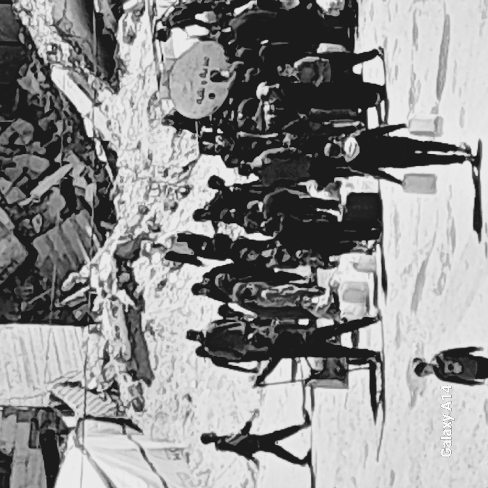 The black and white image, captured in Khan Yunis, Palestinian Territory, depicts a crowded outdoor scene amidst extensive urban destruction. In the background, large piles of concrete rubble from destroyed buildings are prominent, with a white tent structure partially visible on the far left. The midground is dominated by a large gathering of individuals, including adults and children, around a central point. A circular white object bearing Arabic script and numerous white containers on the ground suggest a distribution site. Many individuals within the crowd and in the foreground are seen carrying rectangular jerrycans or similar containers. One person in the foreground walks away from the main group, carrying two jerrycans, while another young individual walks in the bottom-center. An individual on the left side of the frame is seen pulling a cart. The scene is illuminated by daylight, casting distinct shadows. The image exhibits a high-contrast, stylized visual quality.