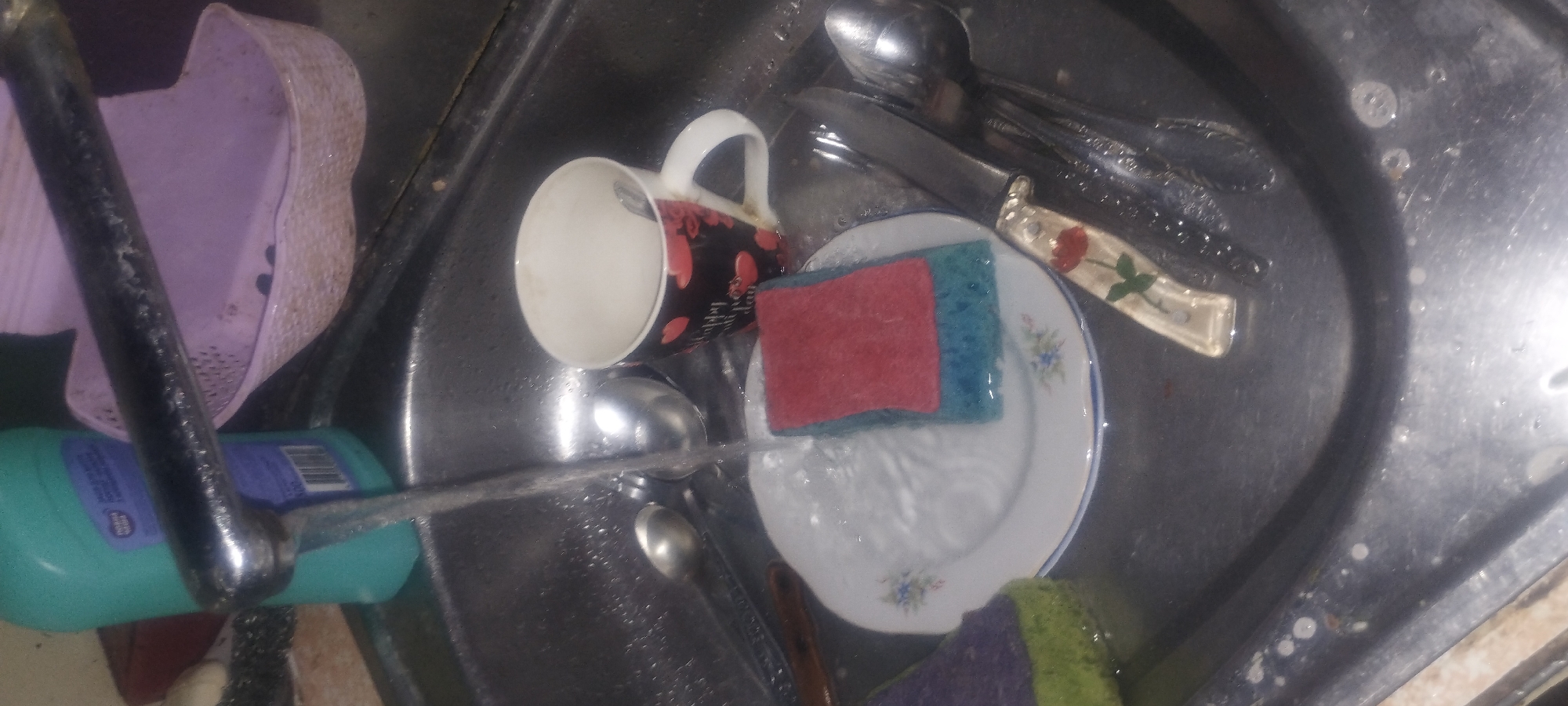 The image depicts a close-up, overhead view of a stainless steel sink in what appears to be a residential kitchen. A stream of water is running from a chrome faucet into the sink. In the sink, there are several items, including a white plate with blue floral trim, a red and blue sponge resting on the plate, a black mug decorated with red hearts and text, a knife with a floral handle, and two spoons. To the left of the sink, a teal-colored bottle of cleaning liquid is visible. In the background, a light purple plastic basket is present. The scene suggests the activity of washing dishes. The location is Zaporizhzhia, Ukraine.