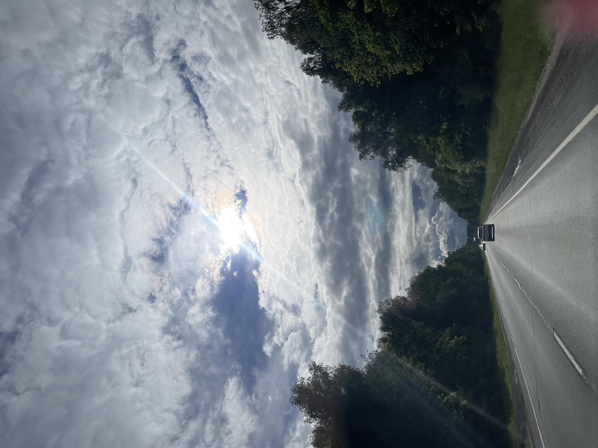 The image depicts a view from a vehicle driving on a paved road. The road extends into the distance with a few cars ahead. Lush green trees line both sides of the road. The sky is filled with clouds, and the sun is visible, creating a bright glare. There is no indication that this image is located in the city of Берегиня Снт, Russia.