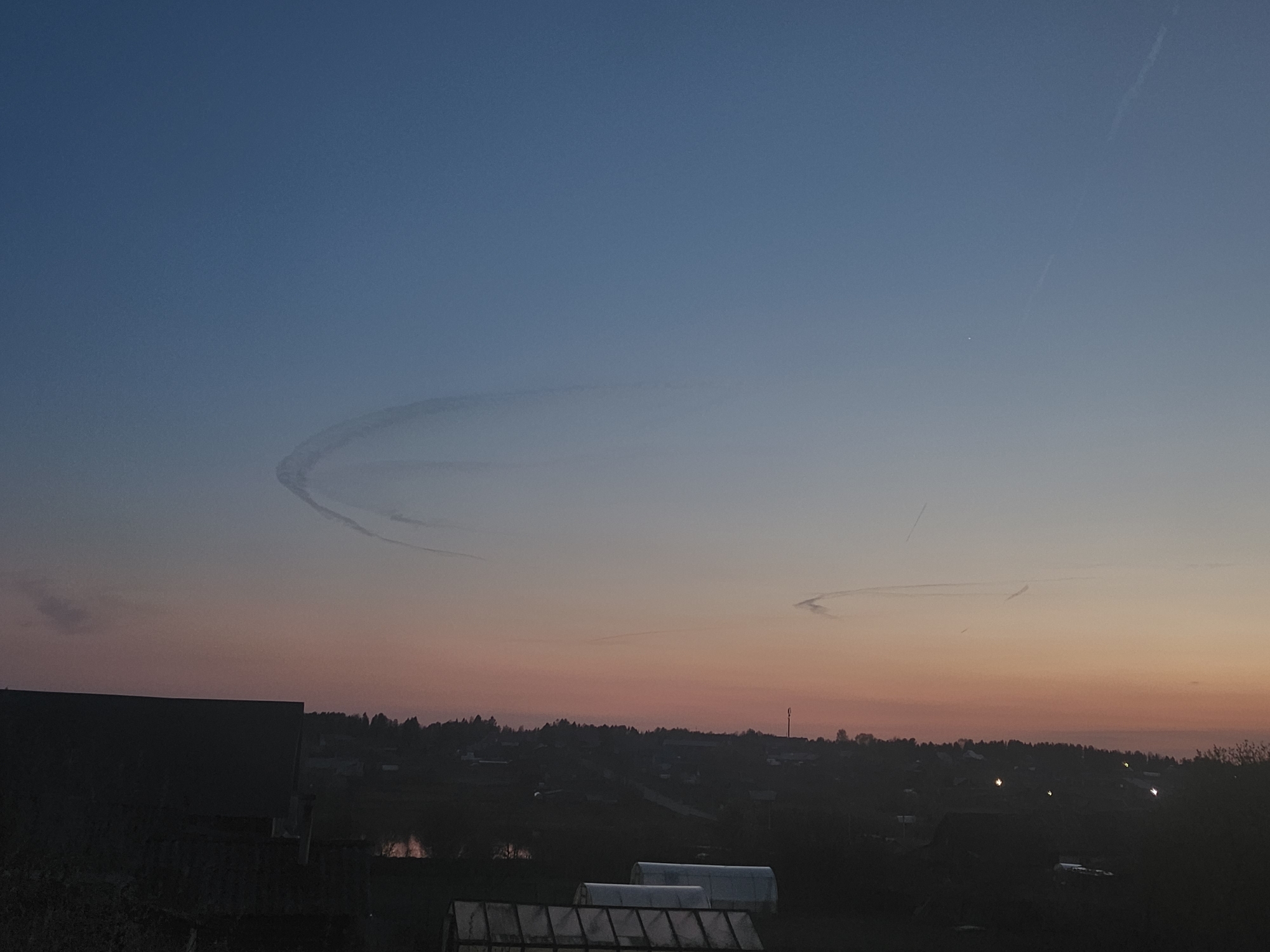 The image captures a dusky evening scene in Krasnogorskoye, Russia. The sky transitions from blue at the top to orange and pink near the horizon. There are visible contrails in the sky, one forming a distinct circular pattern and others appearing as straight lines. The horizon is lined with trees and buildings, appearing as silhouettes. In the foreground, a building with a dark roof is visible on the left. Several greenhouses are also visible. There are a few light points scattered in the landscape, possibly from streetlights or windows. A body of water is partially visible.