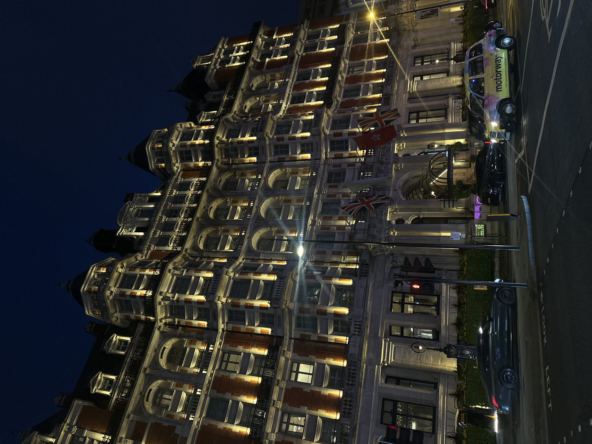 The image shows a street view in London, United Kingdom, at night. On the left, a large, ornate building with many lit windows occupies most of the frame. The architectural style includes red brick accents and arched windows. British and Hong Kong flags are displayed. On the right, a street with visible road markings has a taxi with "motorway" branding, a black car, and a darker-colored car. Traffic lights are present. The sky is dark blue to black.