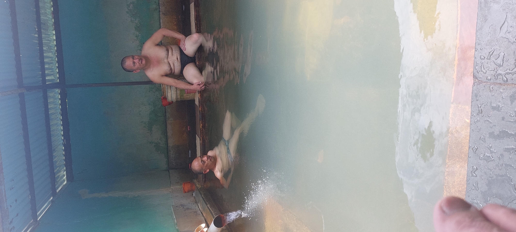 Two men are in a communal outdoor hot spring in Kalath, India. The man on the left is submerged up to his chest in the murky water, leaning back with his eyes closed. He is wearing dark swim trunks. The man on the right is sitting on the edge of the hot spring, also wearing dark swim trunks. He is looking towards the camera with a neutral expression. The hot spring is constructed with concrete walls and a tiled edge. A corrugated metal roof covers part of the structure, supported by metal beams. Several plastic buckets, one red and one white with red trim, are visible near the back wall. Water is flowing into the hot spring from a white pipe on the left side of the frame, creating a splash. The water in the hot spring appears milky and slightly opaque. The surrounding walls are painted a faded teal color, with some signs of wear and discoloration. The floor around the hot spring is wet and appears to be made of large, dark gray tiles. The image is taken from a slightly elevated angle.