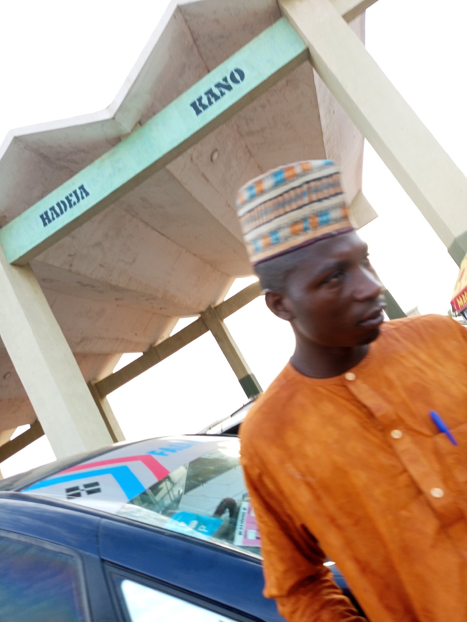 The image captures a street scene in Gombe, Nigeria, under a pedestrian overpass. A young man wearing a colorful, patterned cap and an orange tunic is the central subject, positioned in the foreground and slightly to the right. His head is turned to his right, with his gaze directed outwards. A blue pen is visible in the pocket of his tunic. To the left of the man, the rear of a dark blue car is partially visible. The car's rear windshield displays a graphic with red, blue, and white elements. Above the car and the man, the concrete structure of a pedestrian overpass is prominent. Dark lettering on the overpass reads "HADEJA" on a light green beam on the left, and "KANO" on a similar beam to the right, indicating directional signs or names associated with the location. The overall lighting suggests it is daytime, with bright ambient light.