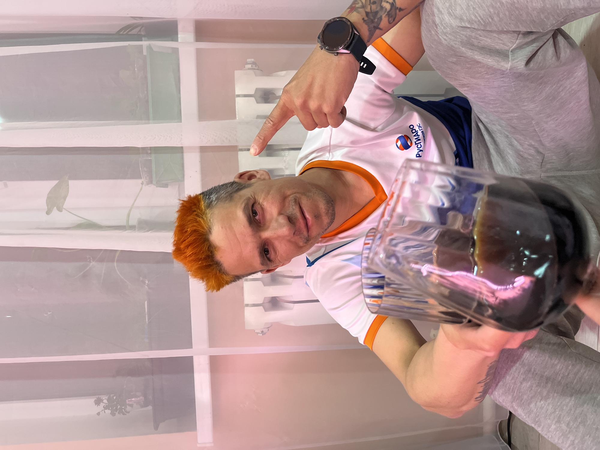 A man with dyed orange hair is indoors, holding a large glass filled with a dark liquid. He is wearing a white t-shirt with orange trim and the word "РУСЧИЧ-МДС" and a logo on the left chest, along with grey sweatpants. He is pointing upwards with his right hand, which also has a black smartwatch on the wrist. The background shows a window with thin white curtains, a white radiator, and a wall that appears to be light pink. The image's location is Chaikovsky, Russia based on the "РУСЧИЧ-МДС" logo, a volleyball club located in that city.