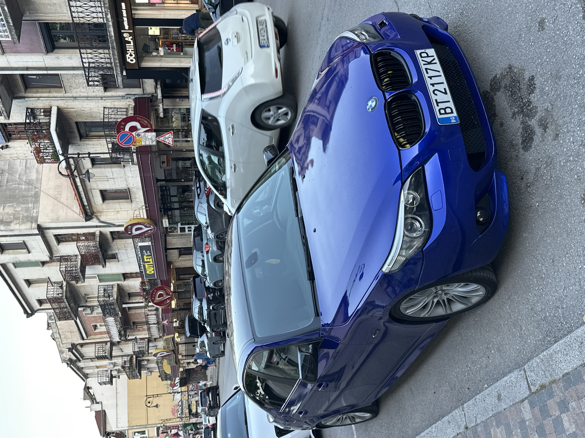 The image depicts a street scene in Veliko Tarnovo, Bulgaria, likely on a pedestrianized or main shopping street given the architecture and storefronts. In the foreground, a vibrant blue BMW sedan is parked, its front license plate clearly visible as "BT 2117 KP". The car is positioned on the right side of the street, facing away from the camera. Further down the street, several other vehicles are visible, including a white Nissan Leaf, a black Audi, and a light blue SUV. The buildings lining the street are multi-story, with a mix of older architectural styles, featuring balconies with intricate metal railings and shops at street level. Signage for businesses like "OUTLET" and "OCHILA optical and sun" are discernible. The sky is overcast, suggesting it might be late afternoon or early evening. A few individuals are walking on the sidewalk in the background, appearing to be pedestrians or shoppers. The overall impression is of a typical urban street in a European city, with the blue BMW as the primary subject of focus.