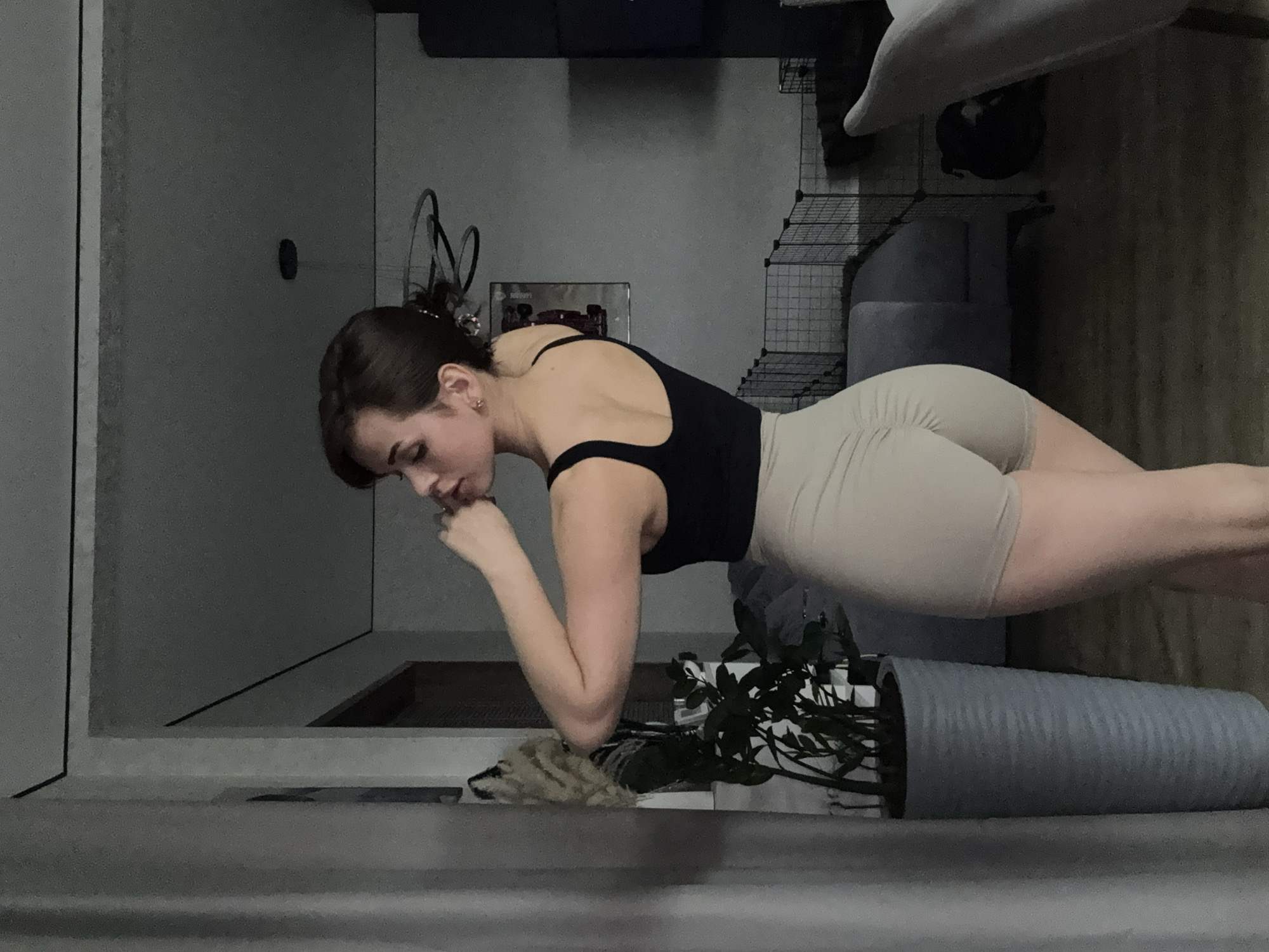 A woman with dark hair and fair skin is doing a plank exercise on the floor of a room. She is wearing a black tank top and tan shorts. Her forearms are resting on the floor, and her head is resting on her hands. To her right is a potted plant in a gray cylindrical pot. Behind her is a wire rack with items on it. The room is dimly lit and appears to be indoors, but the location is unconfirmable from the visual data as Лобаново, Russia.