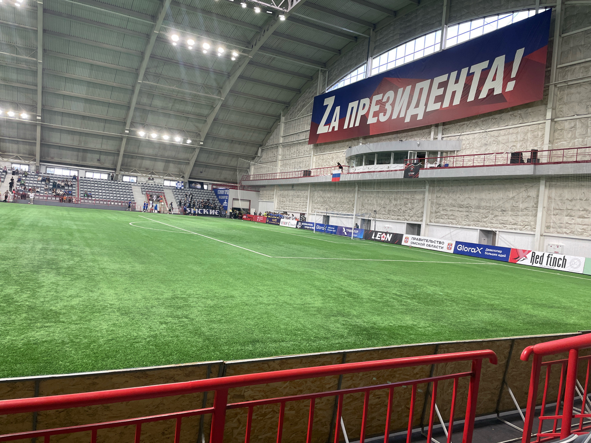 This image shows the interior of an indoor football stadium in Omsk, Russia. The stadium features a bright green artificial turf field with white markings. Spectator stands are visible on the left side of the image, with some people seated. A large banner with Russian text, "ZA ПРЕЗИДЕНТА!", hangs from the ceiling. Several advertisements are displayed along the side of the field, including logos for "LEON," "GloraX," and "Red Finch." A red railing is in the foreground, indicating a spectator area. The overall environment is a well-lit indoor sports facility.