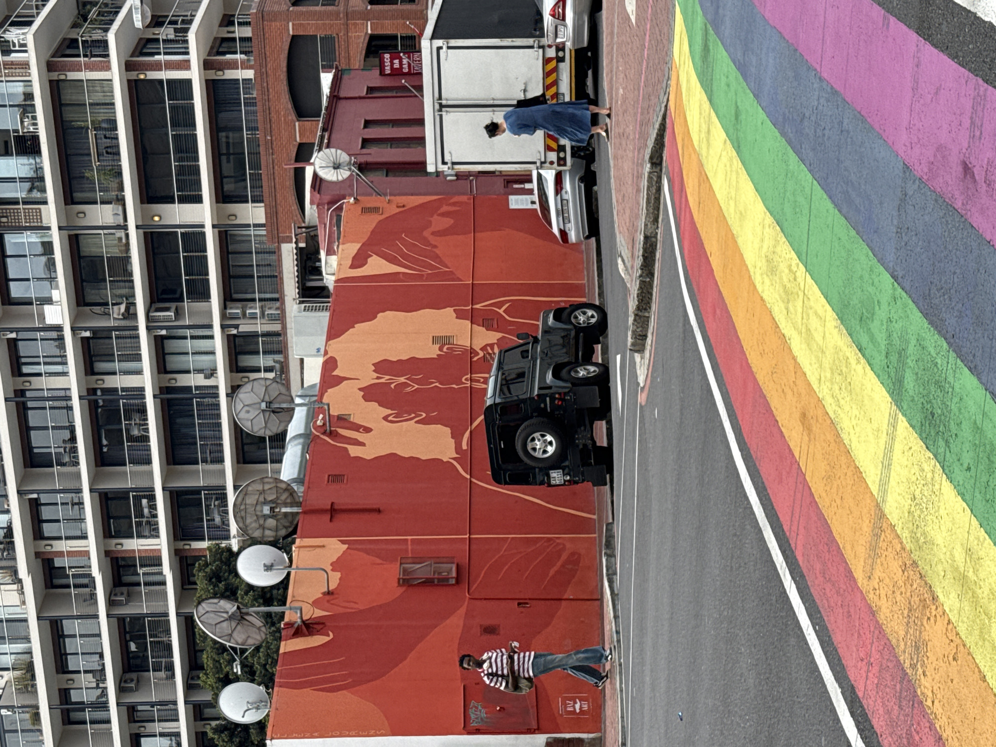 A street in Cape Town, South Africa, features a rainbow crosswalk. A modern apartment building stands tall. A red building with a mural faces the street. A black SUV is parked. A person walks near the mural. A woman in a blue dress walks near a white truck and silver car. Several satellite dishes are mounted on the building.