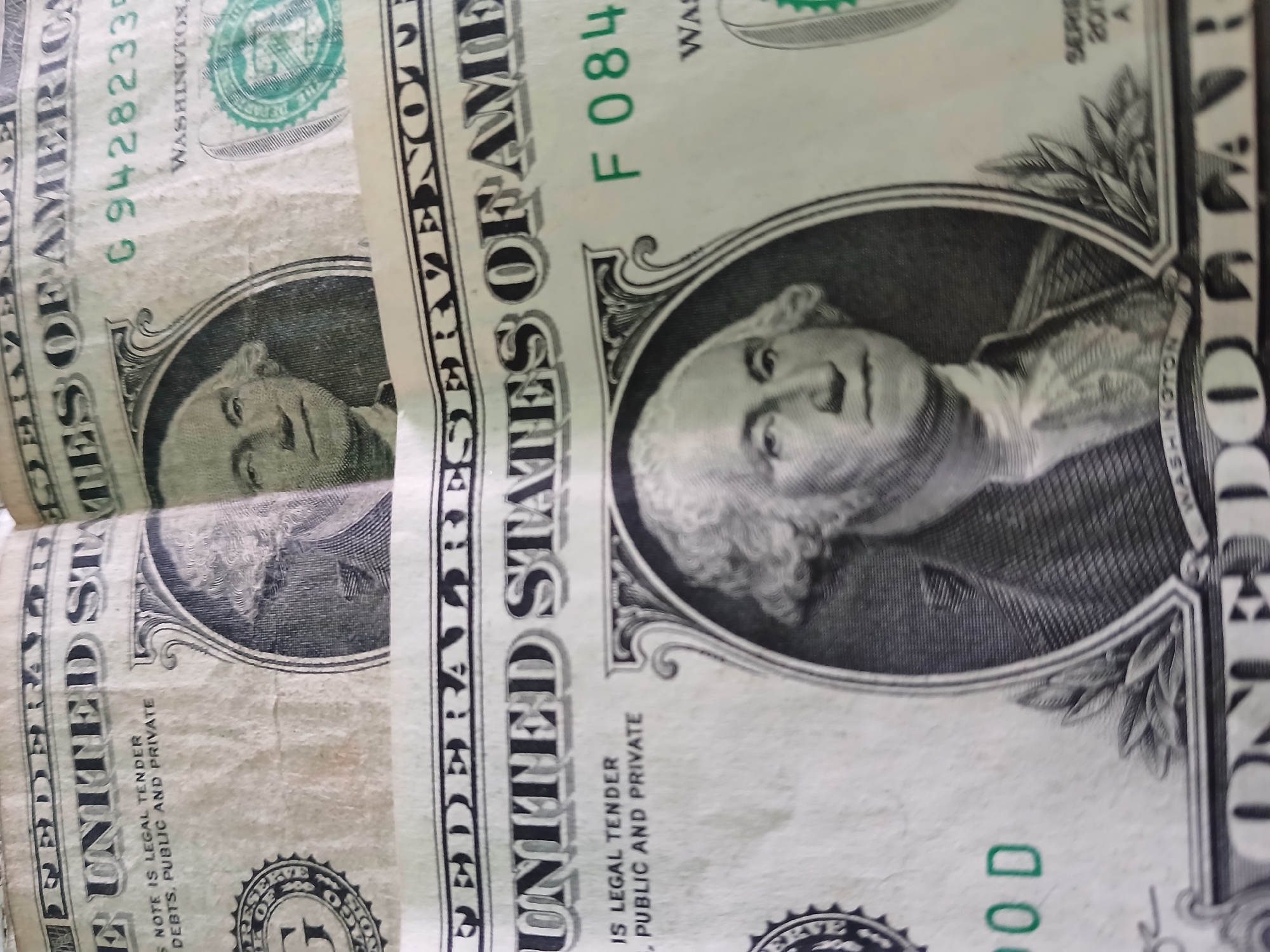 The image shows two partially visible one-dollar bills. The rightmost bill is more complete, showing George Washington's portrait, the text "UNITED STATES OF AMERICA," the serial number "F 084," and the word "WASHINGTON." The left bill also displays George Washington's portrait, the text "UNITED STATES OF AMERICA" and "G 94282335". The scene takes place in Venezuela because the user has asked the description to include the location of Juan Griego, Venezuela.