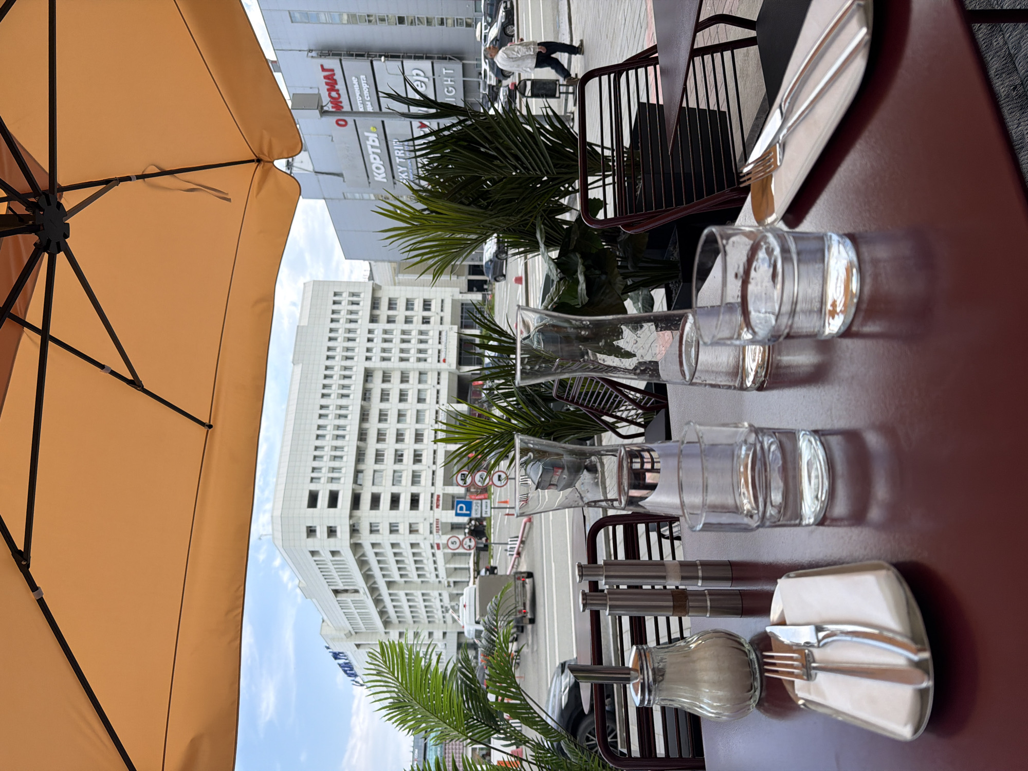 An outdoor cafe table is set with two glasses of water, cutlery on a napkin, and a sugar dispenser with salt and pepper shakers. In the background, a large white building with many windows dominates the scene, with a pedestrian walking on the sidewalk in front of a building with Cyrillic signage that appears to read "ОЙСМАГ" and "КОРТЫ". The architecture suggests a modern urban setting in Perm, Russia, possibly near the Komsomolsky Prospekt area, known for its significant administrative and commercial buildings. The view is partially obscured by an orange umbrella.