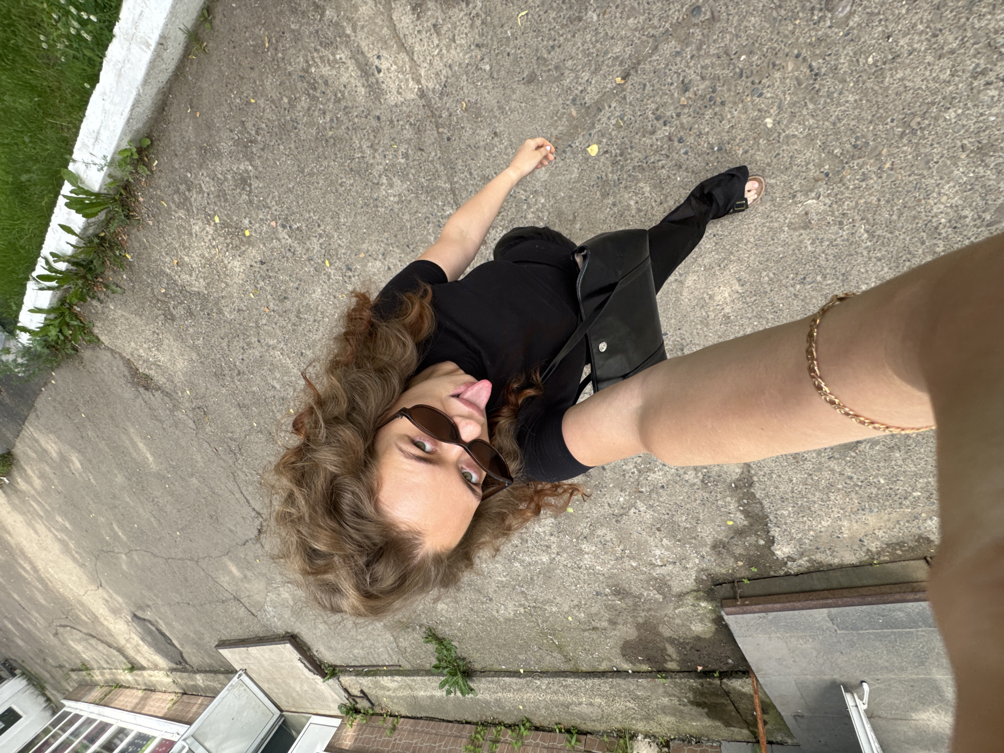 A young woman with curly brown hair and sunglasses is standing on a concrete path. She is wearing a black t-shirt and black pants, and carrying a black purse. She is sticking her tongue out at the camera. The background shows a building with large windows and some greenery. The scene appears to be in an urban environment in Chaikovsky, Russia.