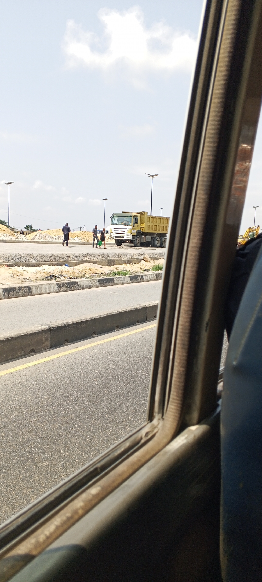 An external view from inside a vehicle shows a construction site with a yellow dump truck parked on a dirt road. Three men are visible near the truck. One man in a dark blue shirt and pants walks away from the truck. Two other men, one in a dark t-shirt and the other in a lighter shirt, stand together to the left of the dump truck. A yellow excavator is partially visible in the background to the right. Several tall, modern streetlights are positioned throughout the scene. The ground is covered with sand and construction debris. The sky is bright and partly cloudy. The foreground of the image is dominated by the interior of the vehicle, with a dark blue seat and a metal window frame.